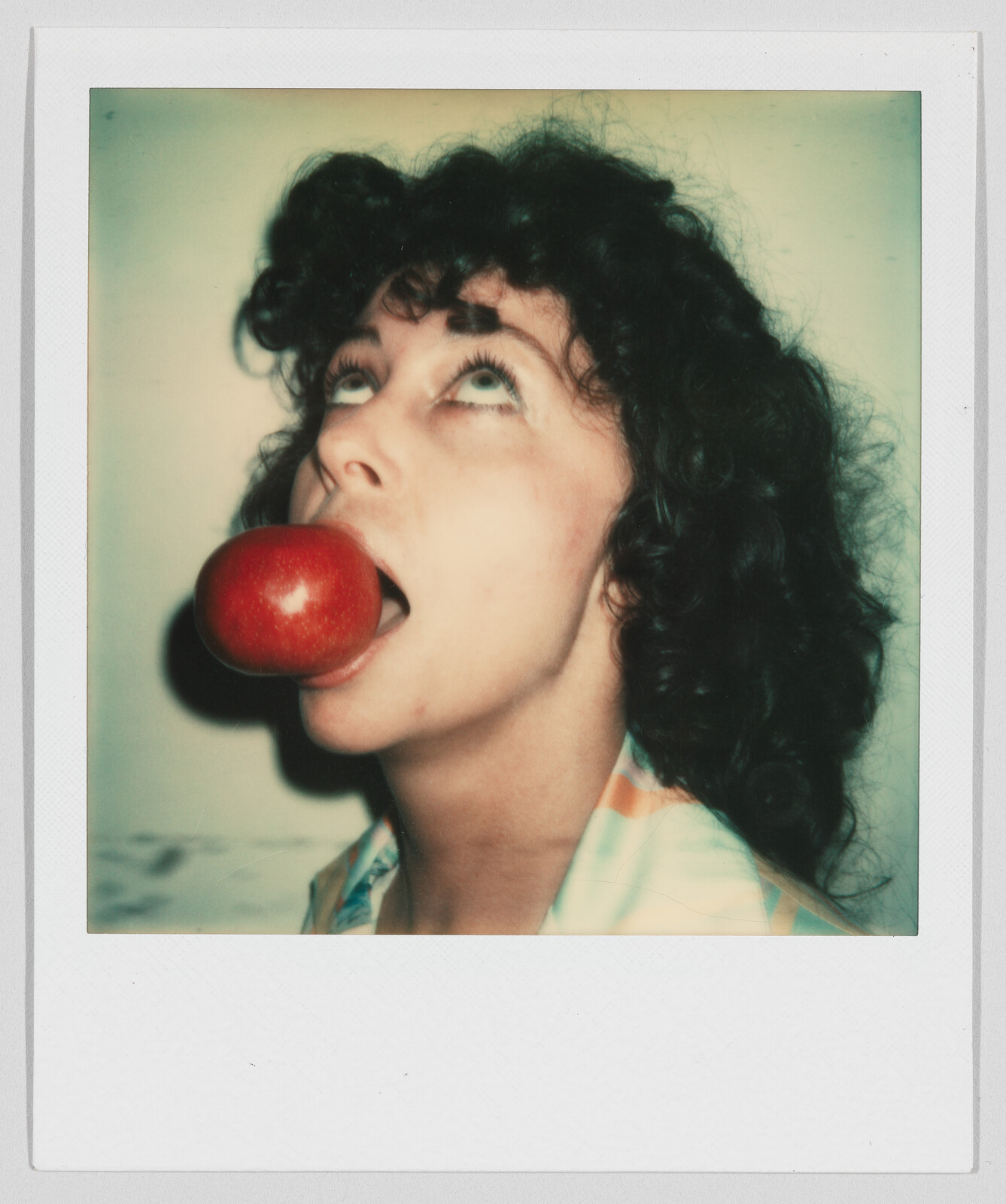 Person with curly hair looking up, holding a large red apple in their open mouth.