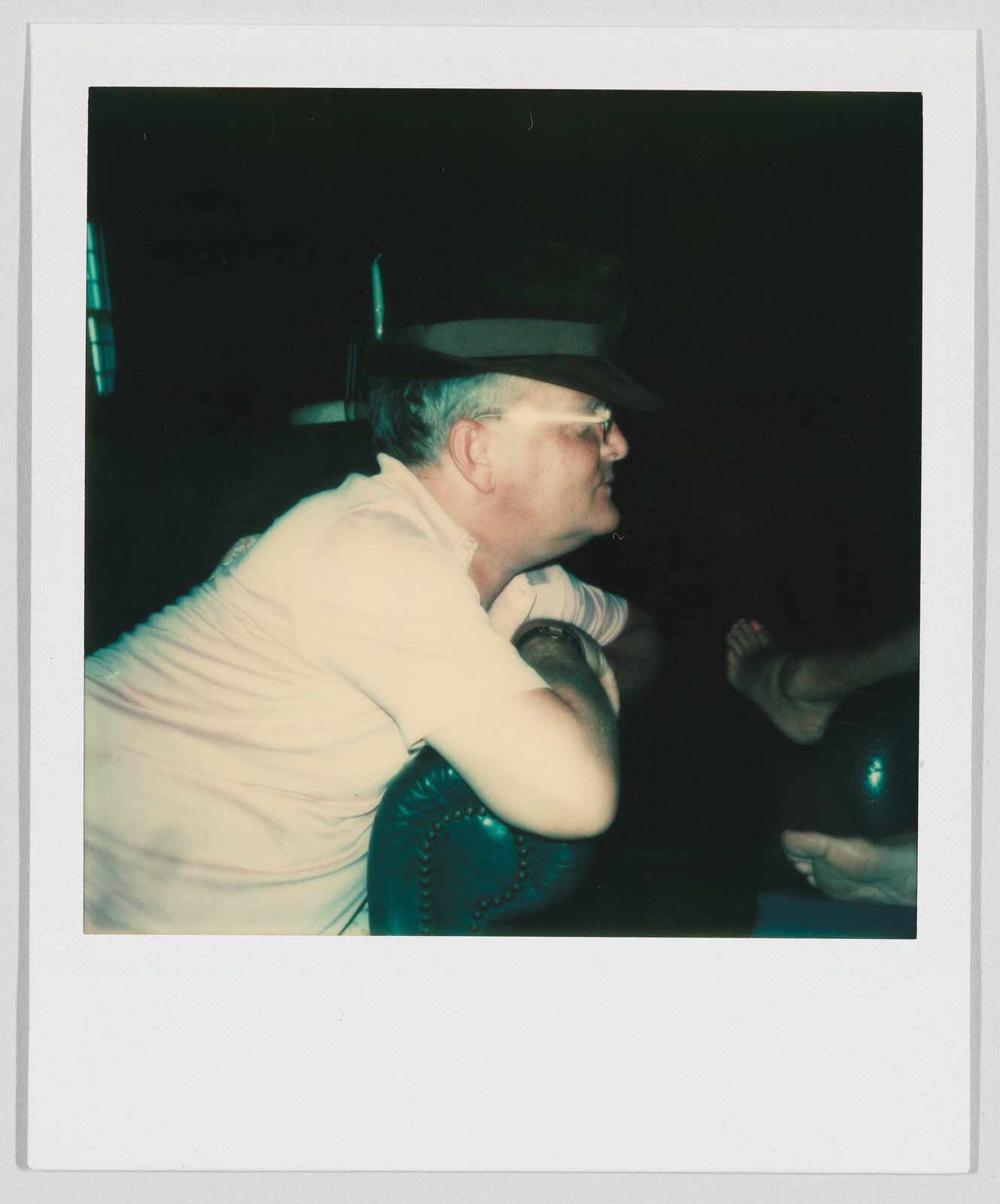 Man wearing glasses and a hat leans forward on a chair, looking to the right in a dim room.
