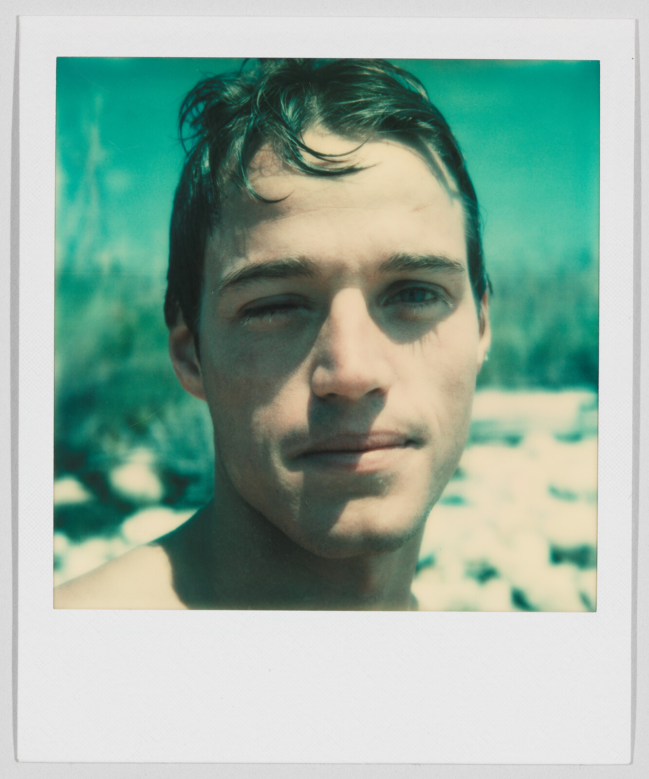 A young man with wet hair looks at the camera outdoors, with a blurred background of sky and trees.