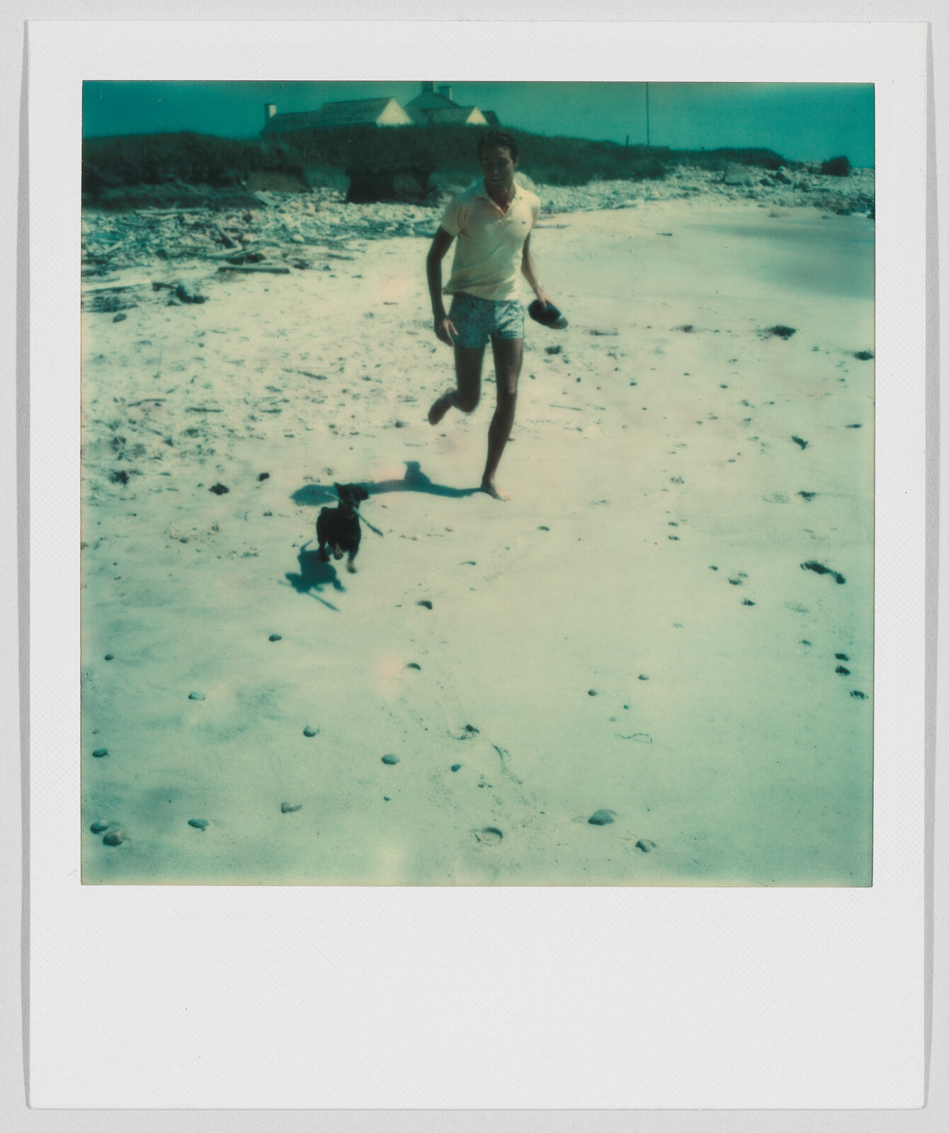 A man runs barefoot on a sandy beach with a small black dog beside him.