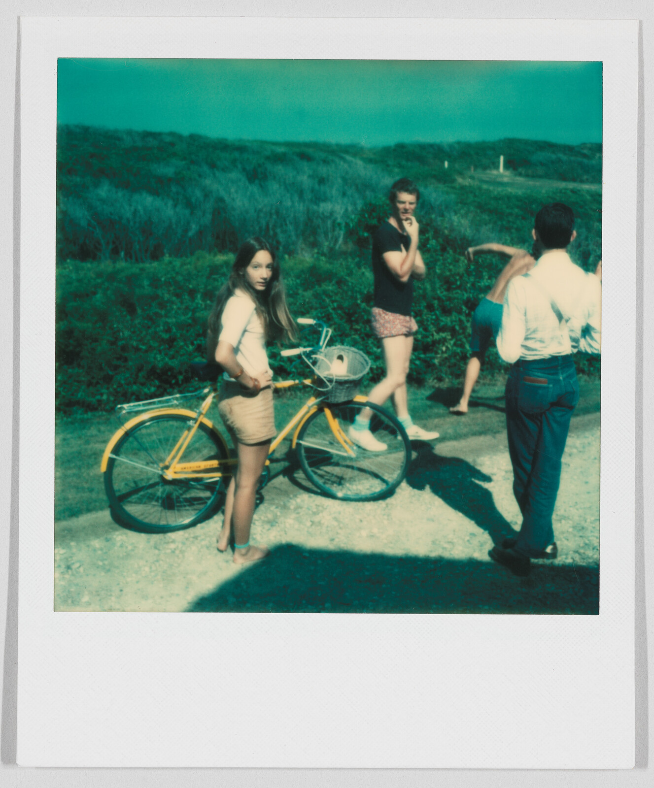 Four people stand on a path near bushes; one young woman holds a yellow bicycle and looks toward the camera.
