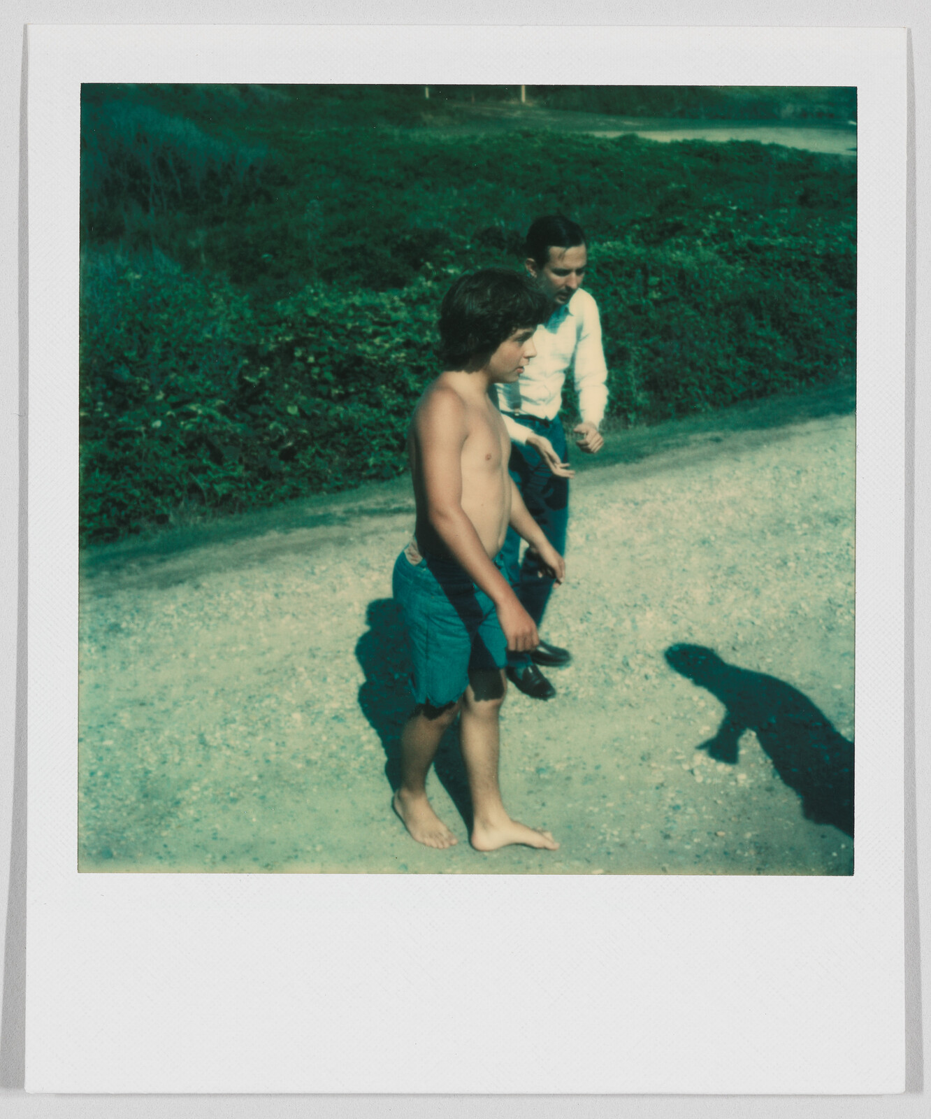 A shirtless boy and a man walk on a dirt path, with greenery in the background.