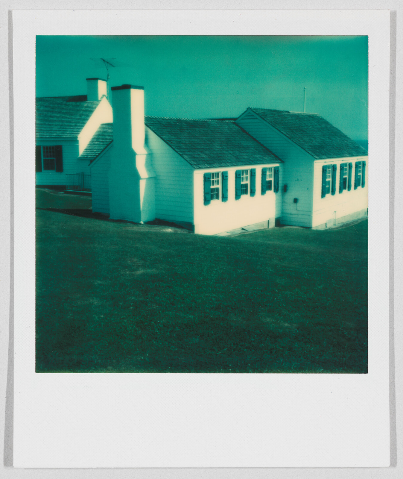 A small white house with dark shutters and a chimney sits on a grassy lawn under a clear sky.