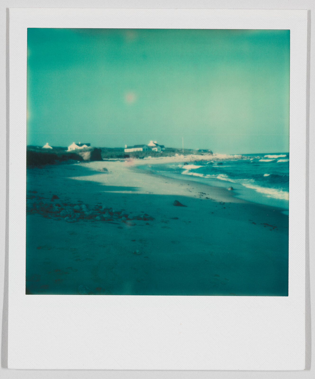 Sandy beach with gentle waves, a few scattered rocks, and small houses in the distance under a clear sky.