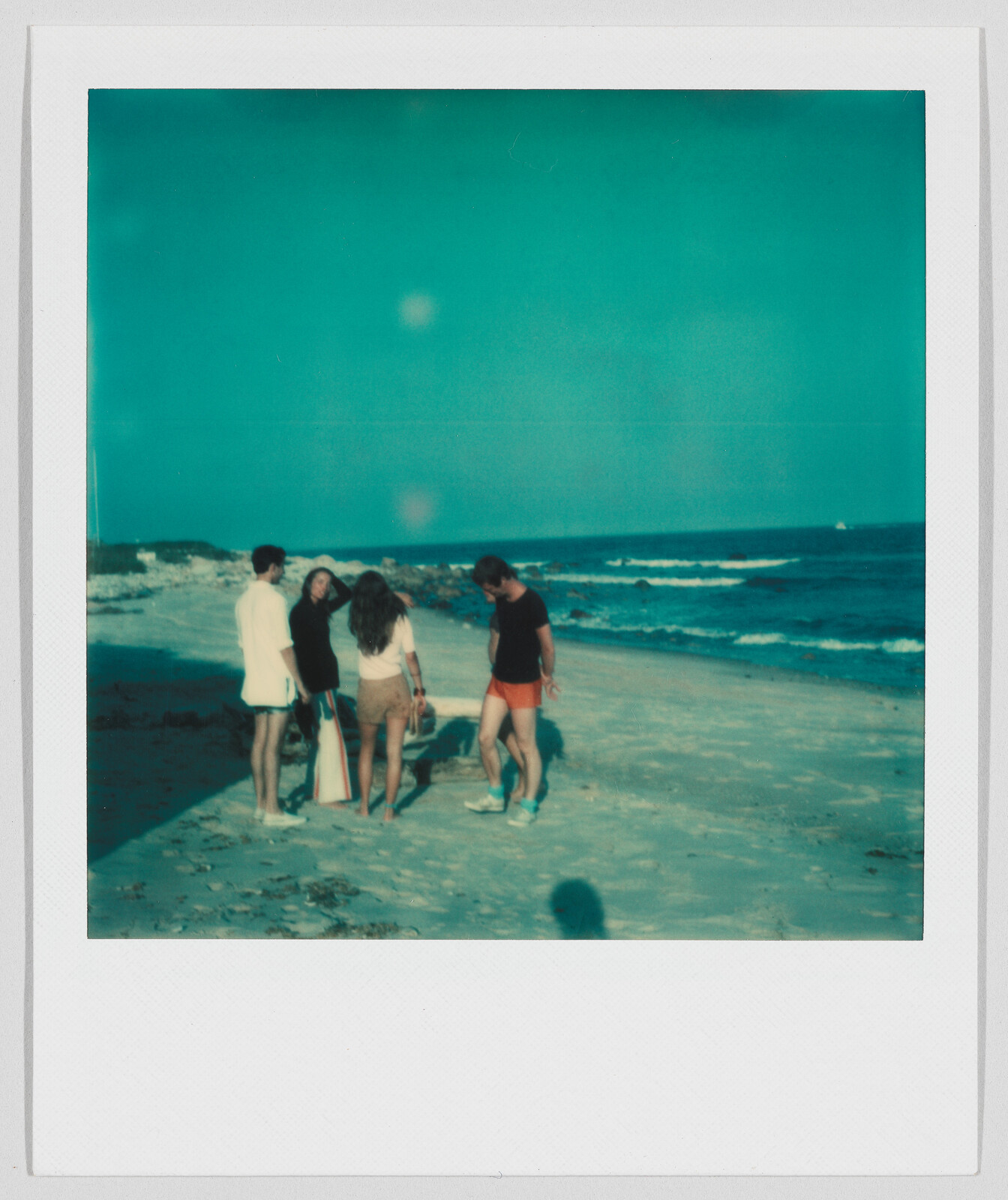 Four people stand together on a sandy beach near the ocean, talking and enjoying the sunny weather.