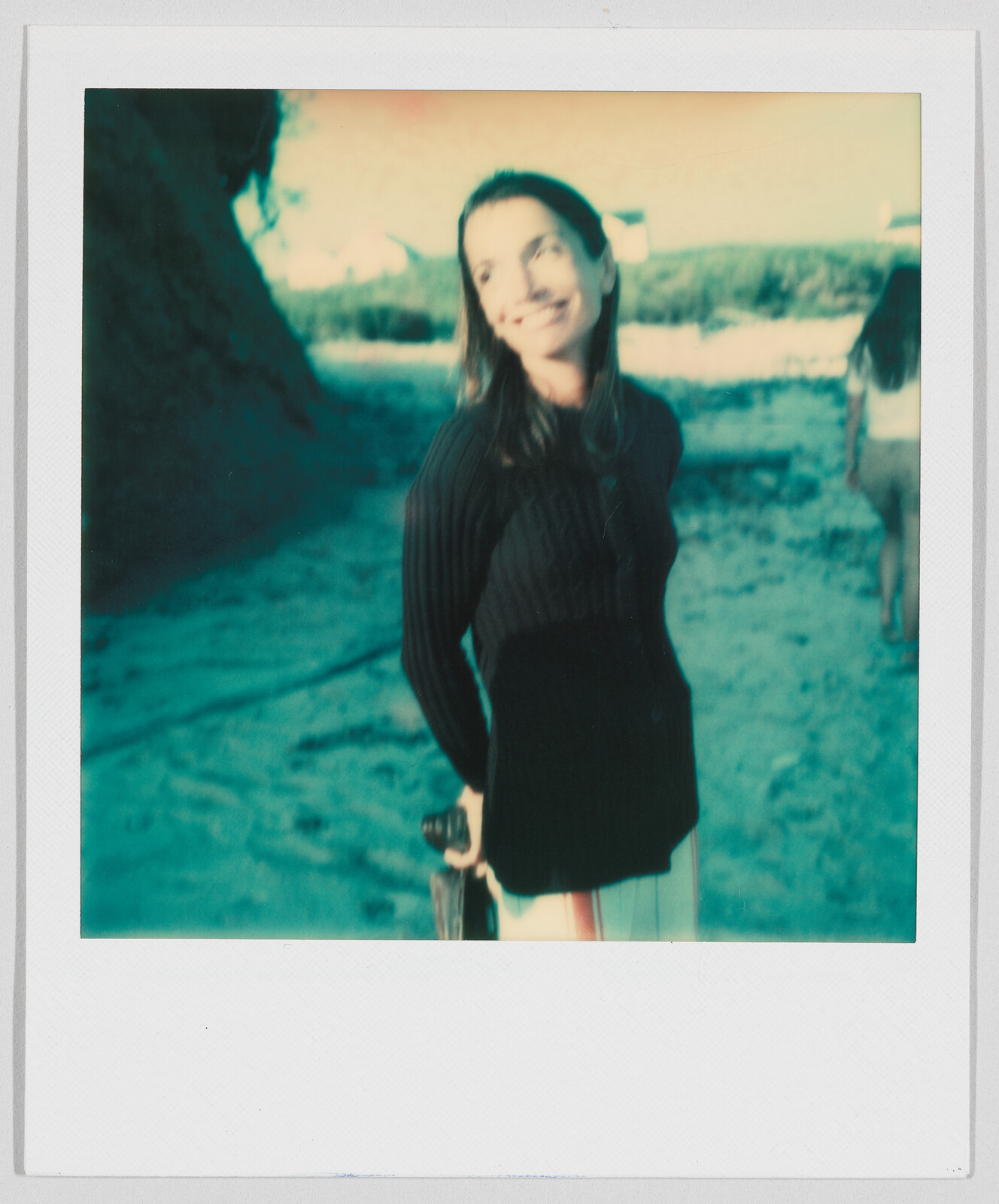 Woman smiling and holding a skateboard on a sandy beach, with another person walking in the background.
