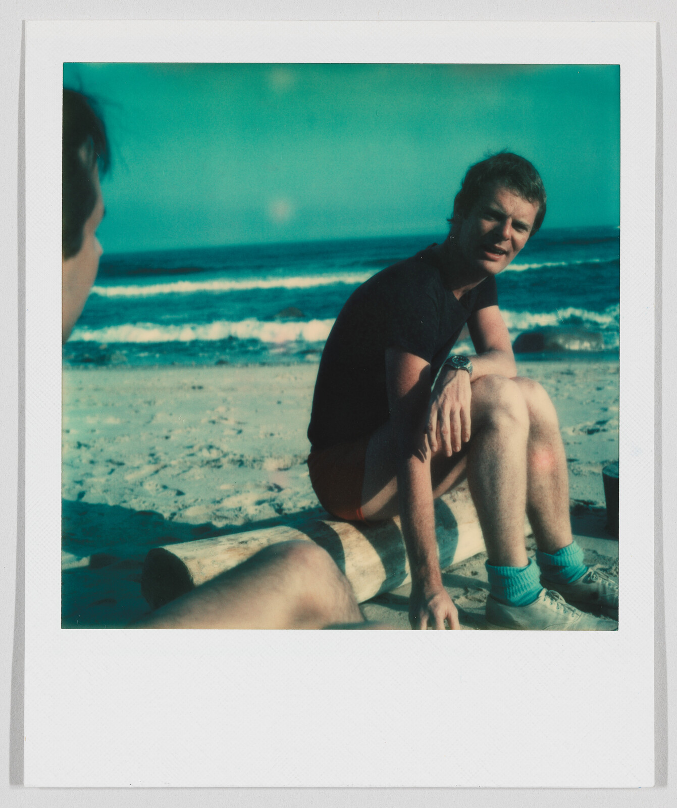 A man sits on a log at the beach, looking toward another person, with waves in the background.