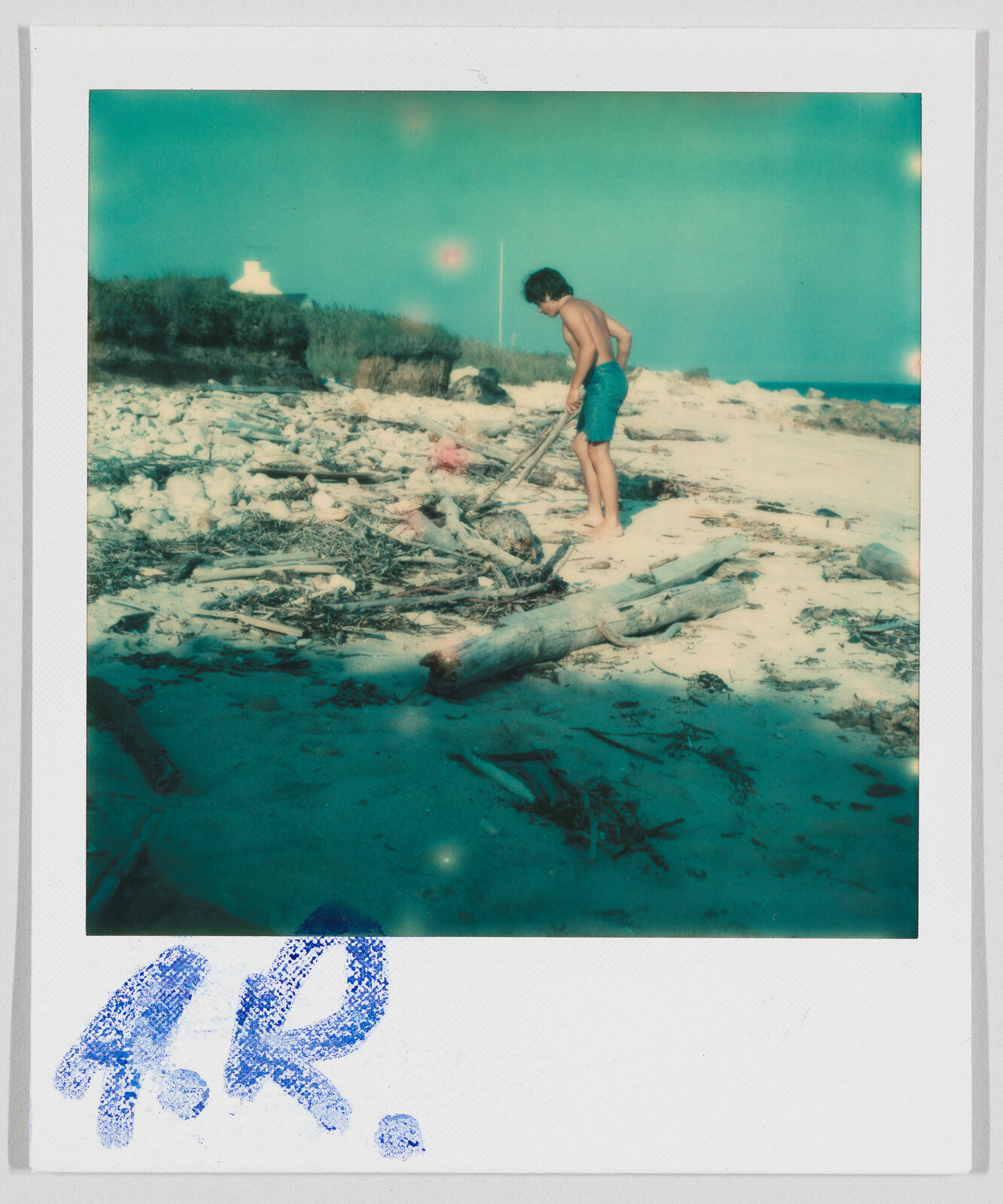 A boy in blue shorts stands on a sandy beach, picking up driftwood. The initials "A.R." appear below.