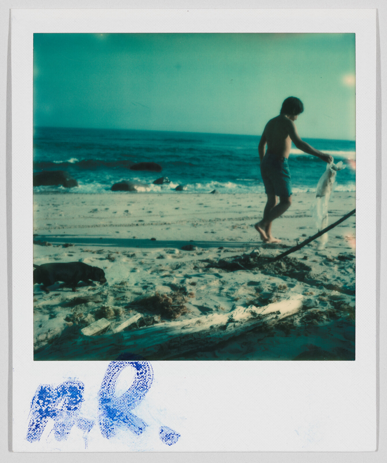 A person in swim trunks walks barefoot on a sandy beach near the ocean, holding a towel.