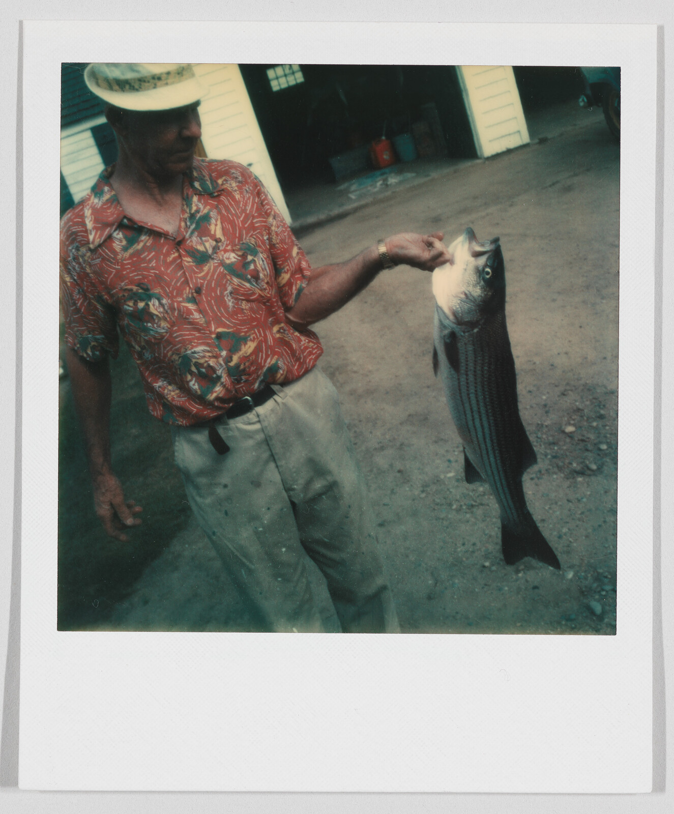 Man in a patterned shirt and hat holding a large fish by the mouth outside a building.