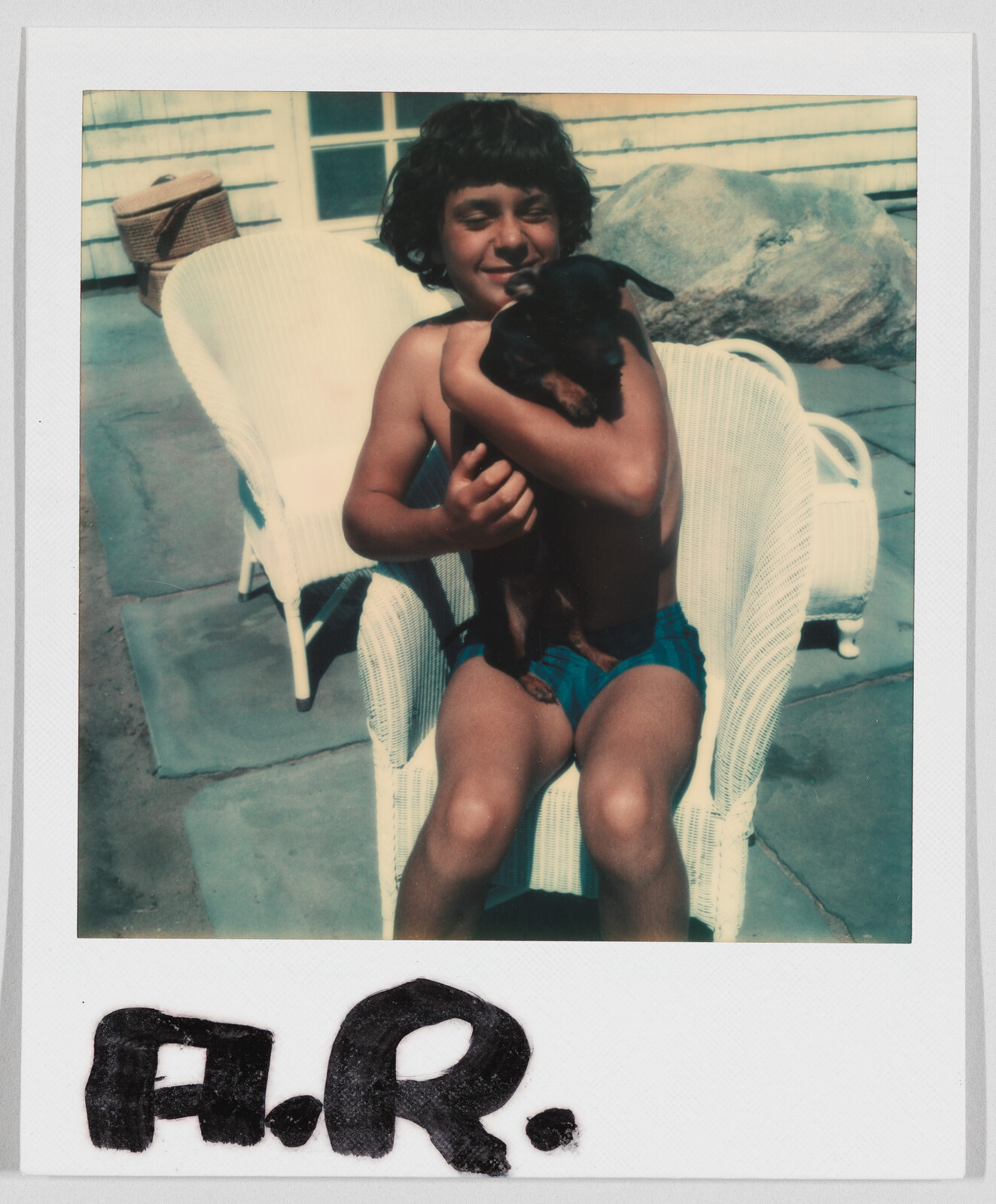 A boy in swim trunks sits on a white chair outside, hugging a small black dog. "A.R." is written below.