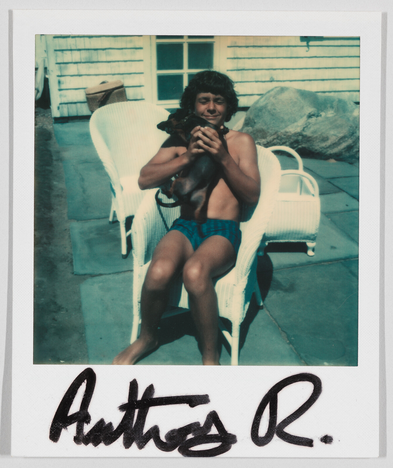 A boy in swim trunks sits on a white chair outside, holding a small dog. Signature reads "Anthos R."