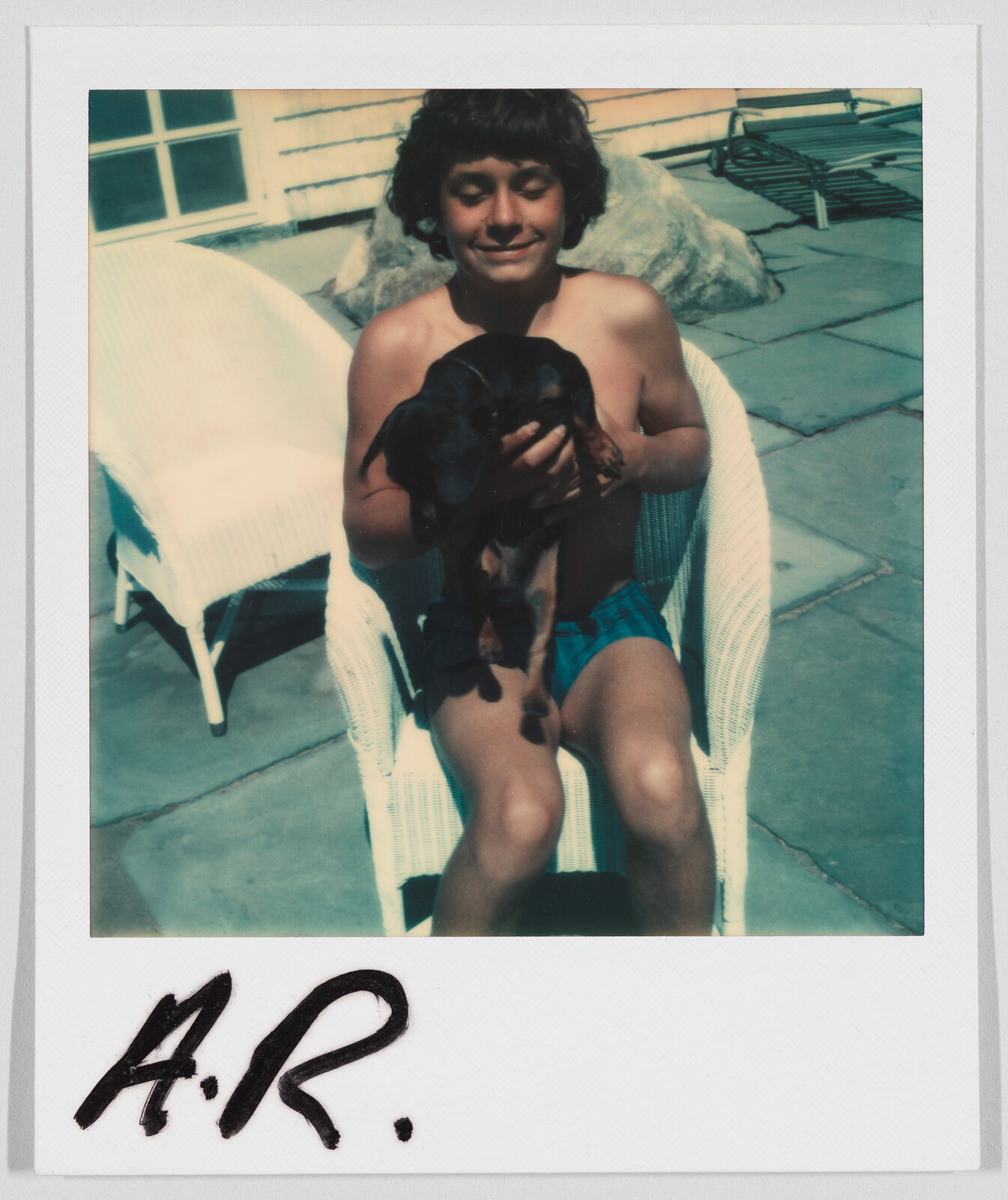 A smiling child in swim trunks sits on a white chair outdoors, holding a small black dog. "A.R." is written below.
