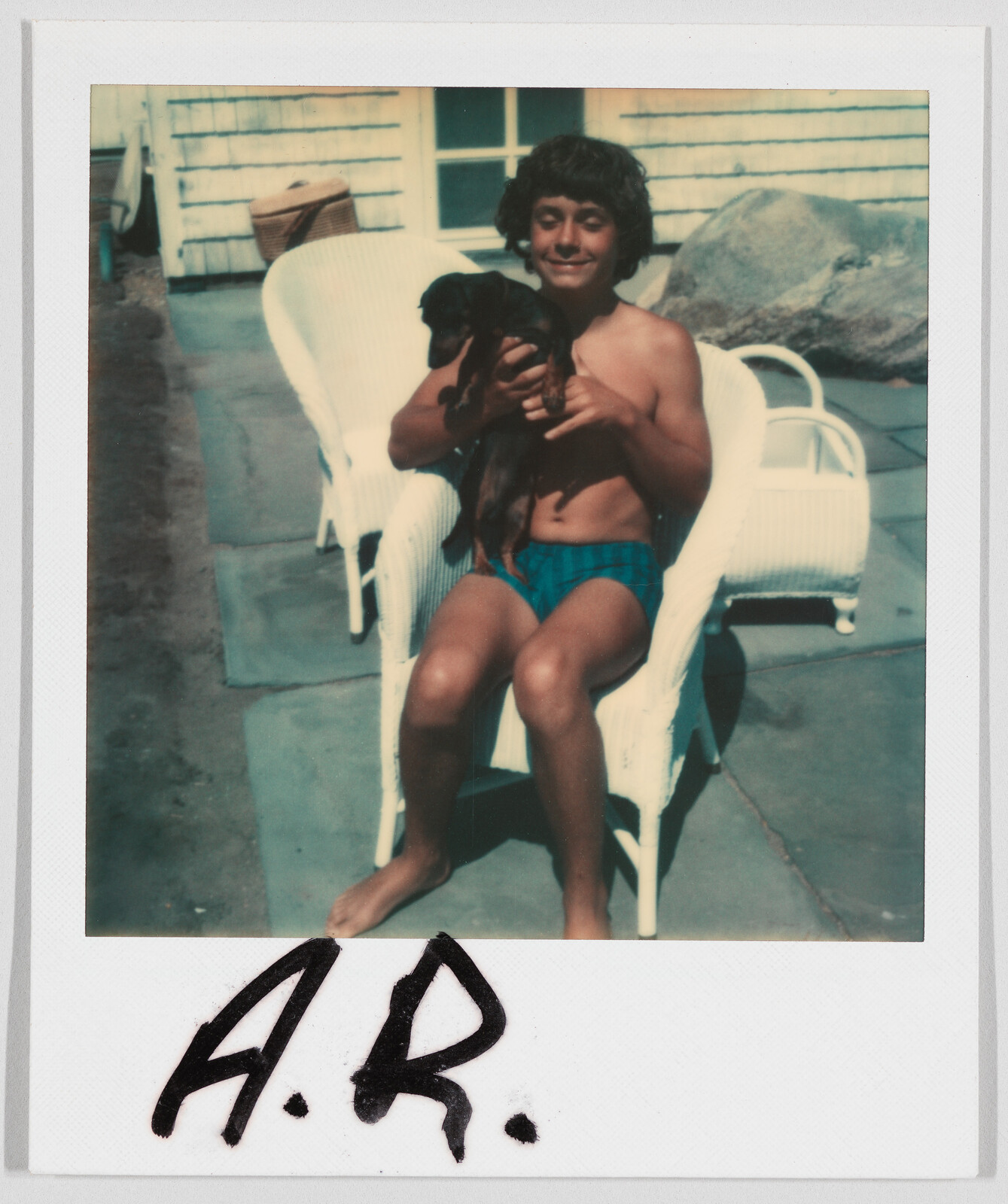 A boy in swim trunks sits on a white chair outside, smiling and holding a small black dog. The initials "A.R." are written at the bottom.