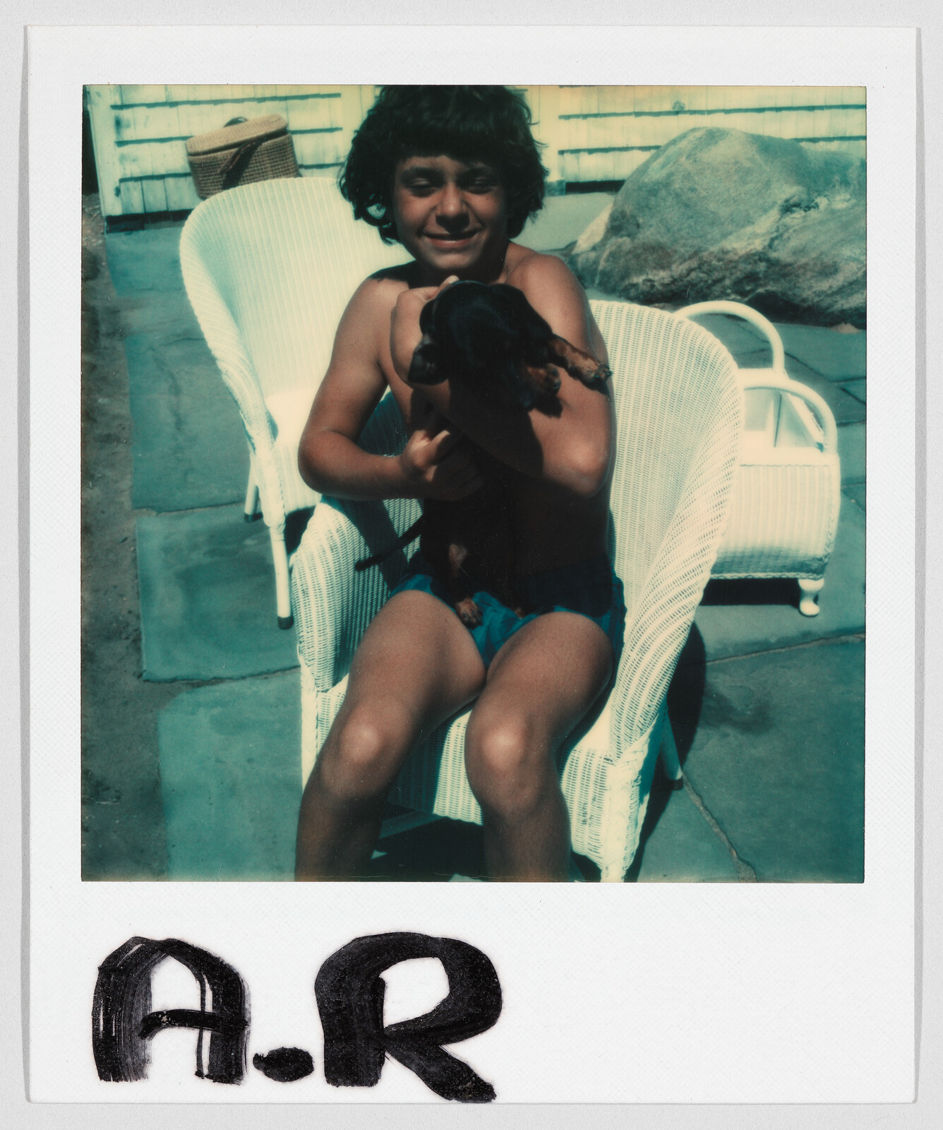 A boy in swim trunks sits on a white chair outside, holding a small black and brown dog. The letters "A.R" are written at the bottom.