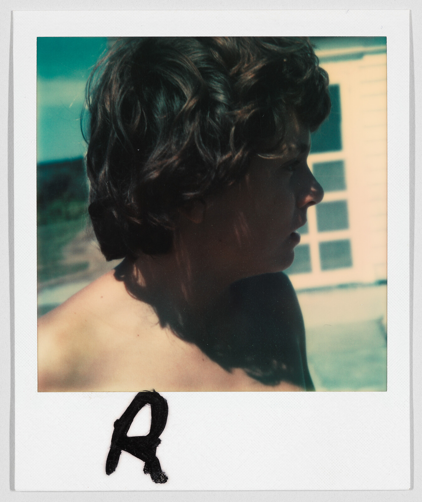 A person with curly hair looks to the side outdoors, with a large letter "R" written below.