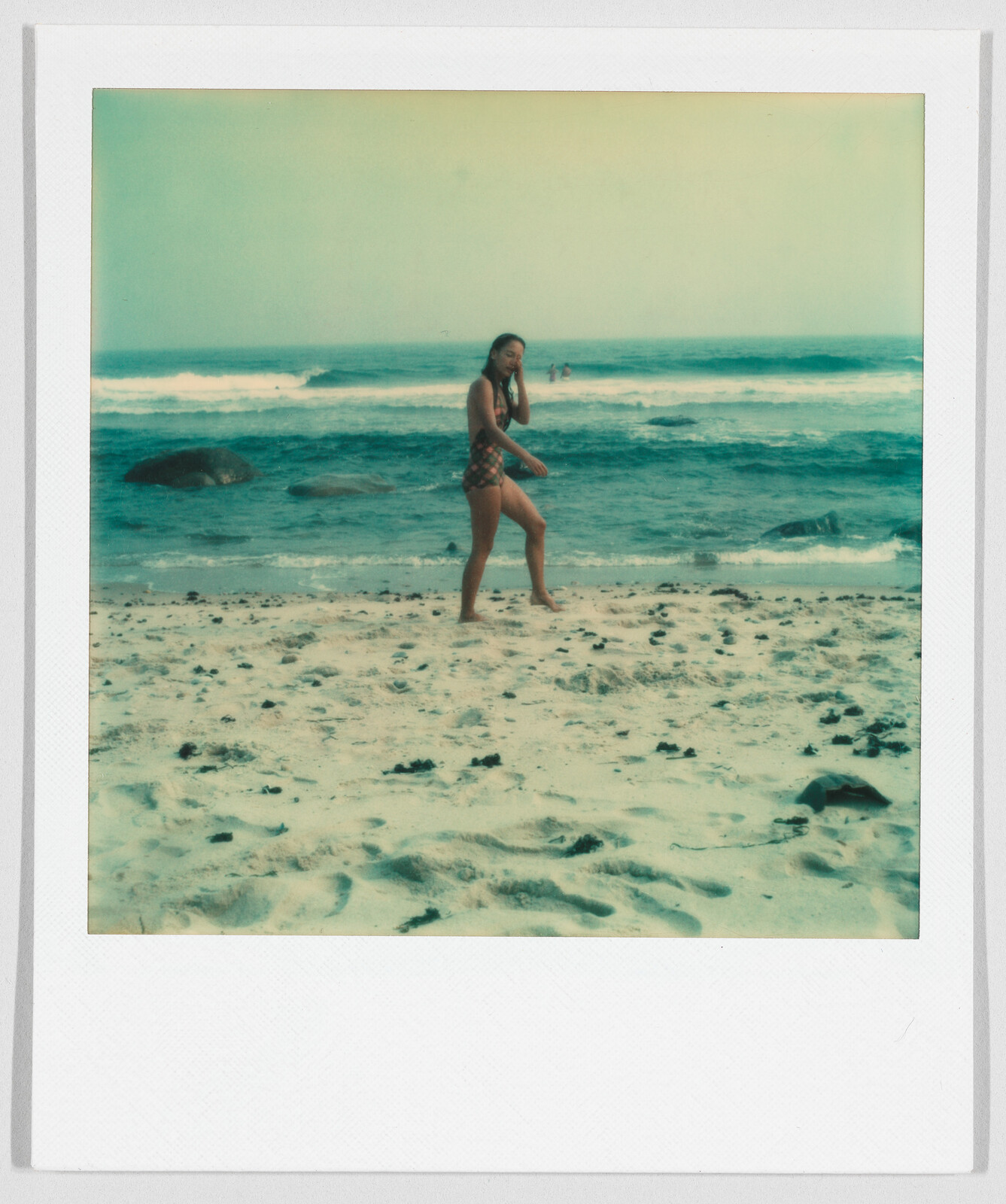A woman in a swimsuit stands on a sandy beach near the ocean, with waves and rocks behind her.