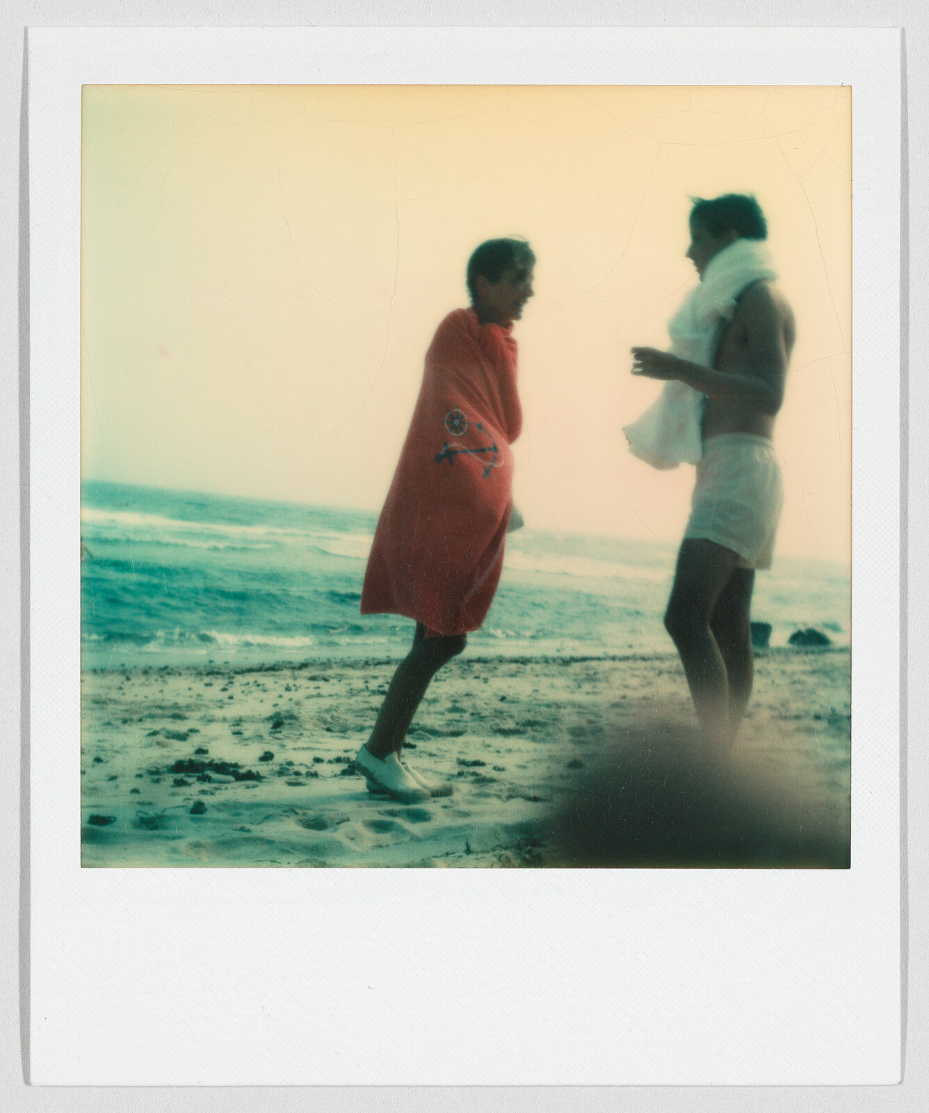 Two people stand on a sandy beach, both wrapped in towels, with waves in the background.