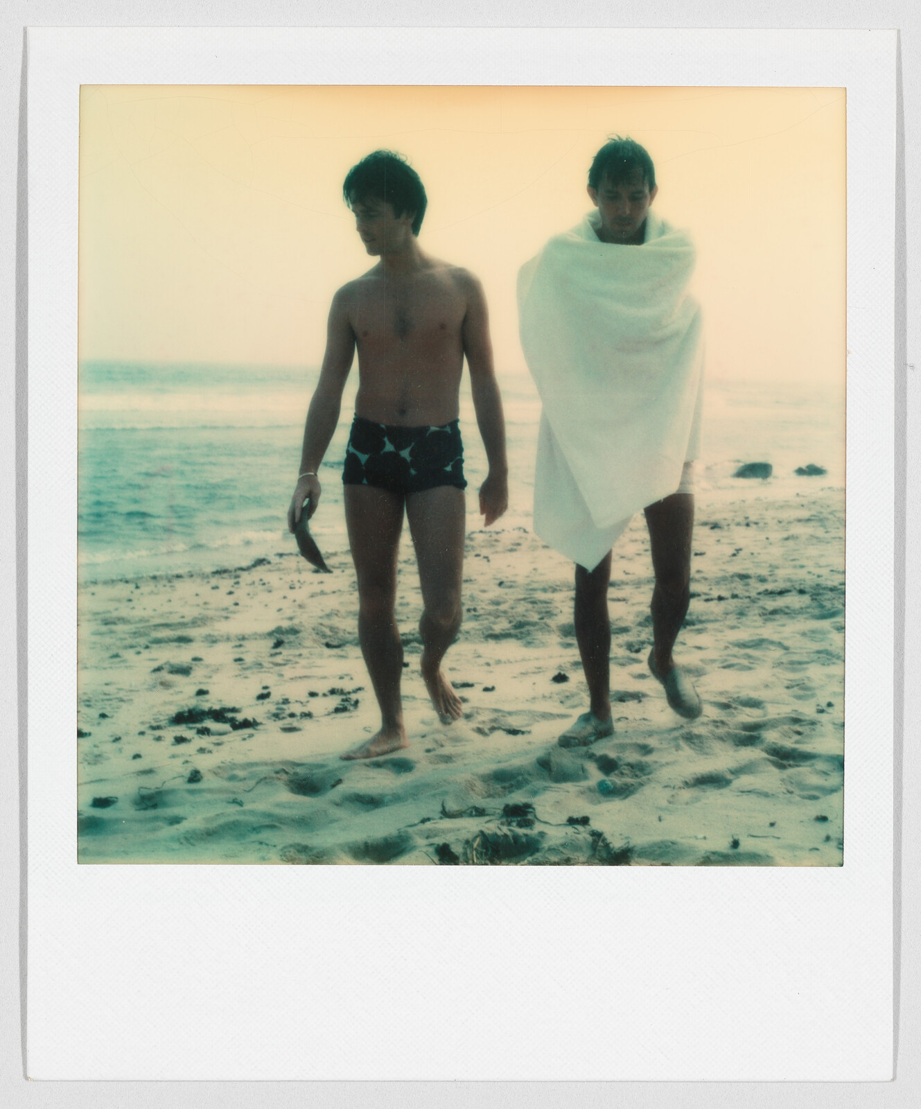 Two young men walk on a sandy beach, one in swim trunks and the other wrapped in a towel.