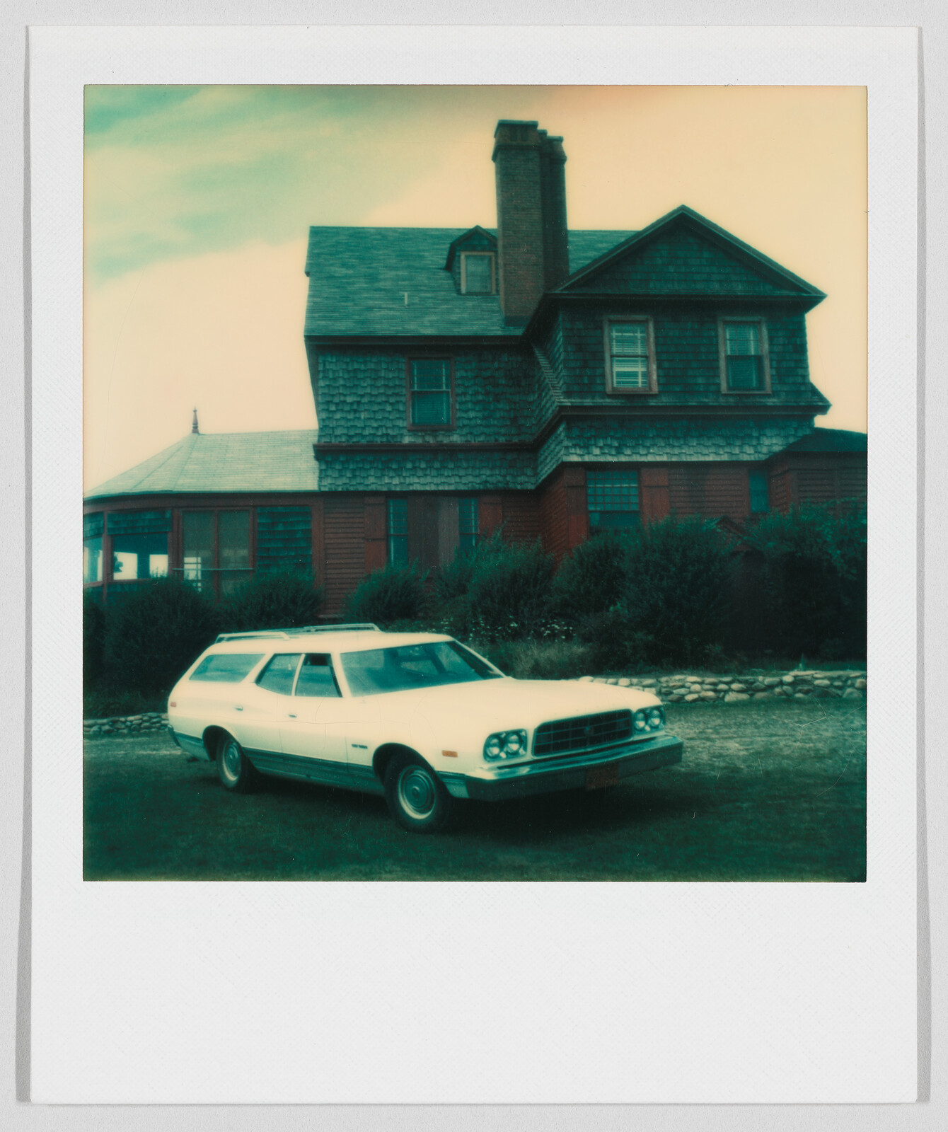 A white station wagon is parked on grass in front of a large, old wooden house with a chimney.