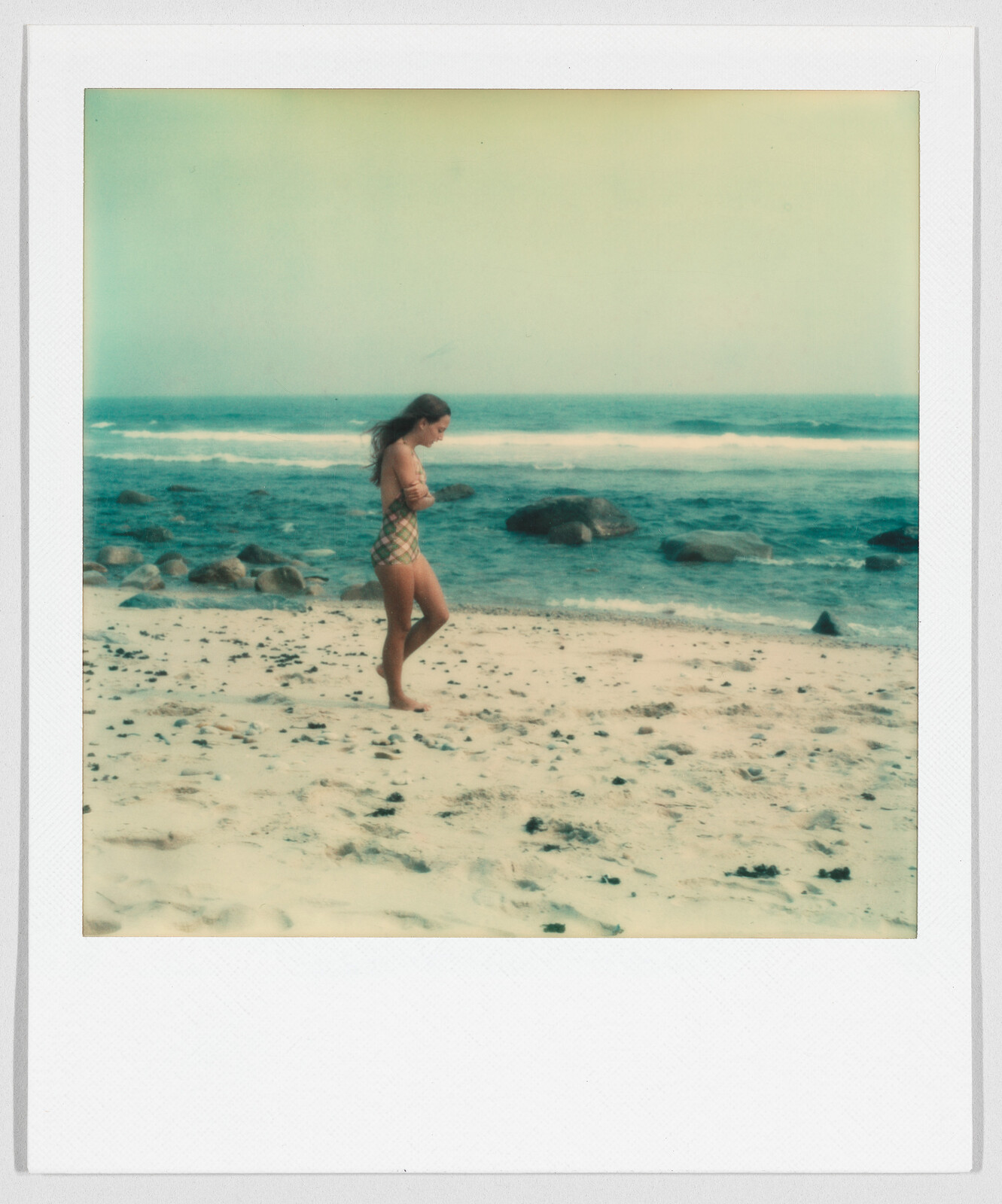 A woman in a swimsuit walks alone on a sandy beach near the ocean, with rocks in the background.