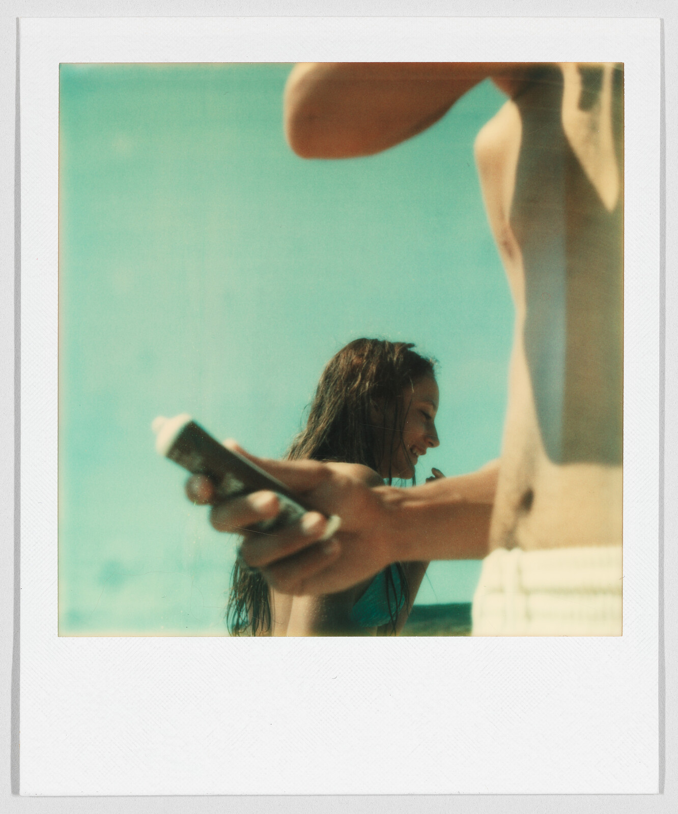 Two people at the beach, one holding sunscreen, with a clear blue sky in the background.