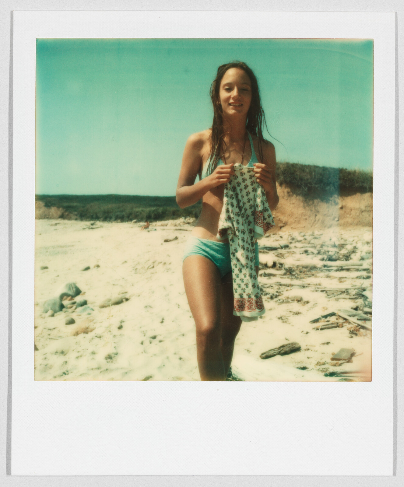 A woman in a swimsuit stands on a sandy beach, holding a patterned shirt and smiling.