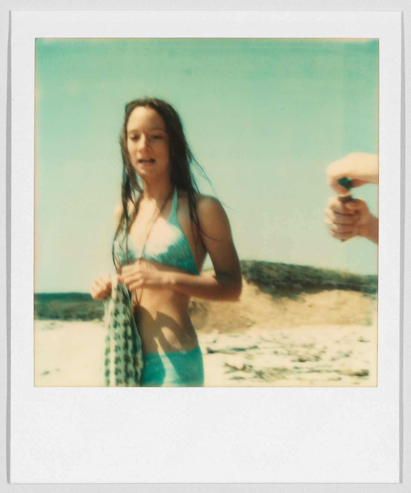 A young woman in a blue bikini stands on a sandy beach, holding a patterned cloth.