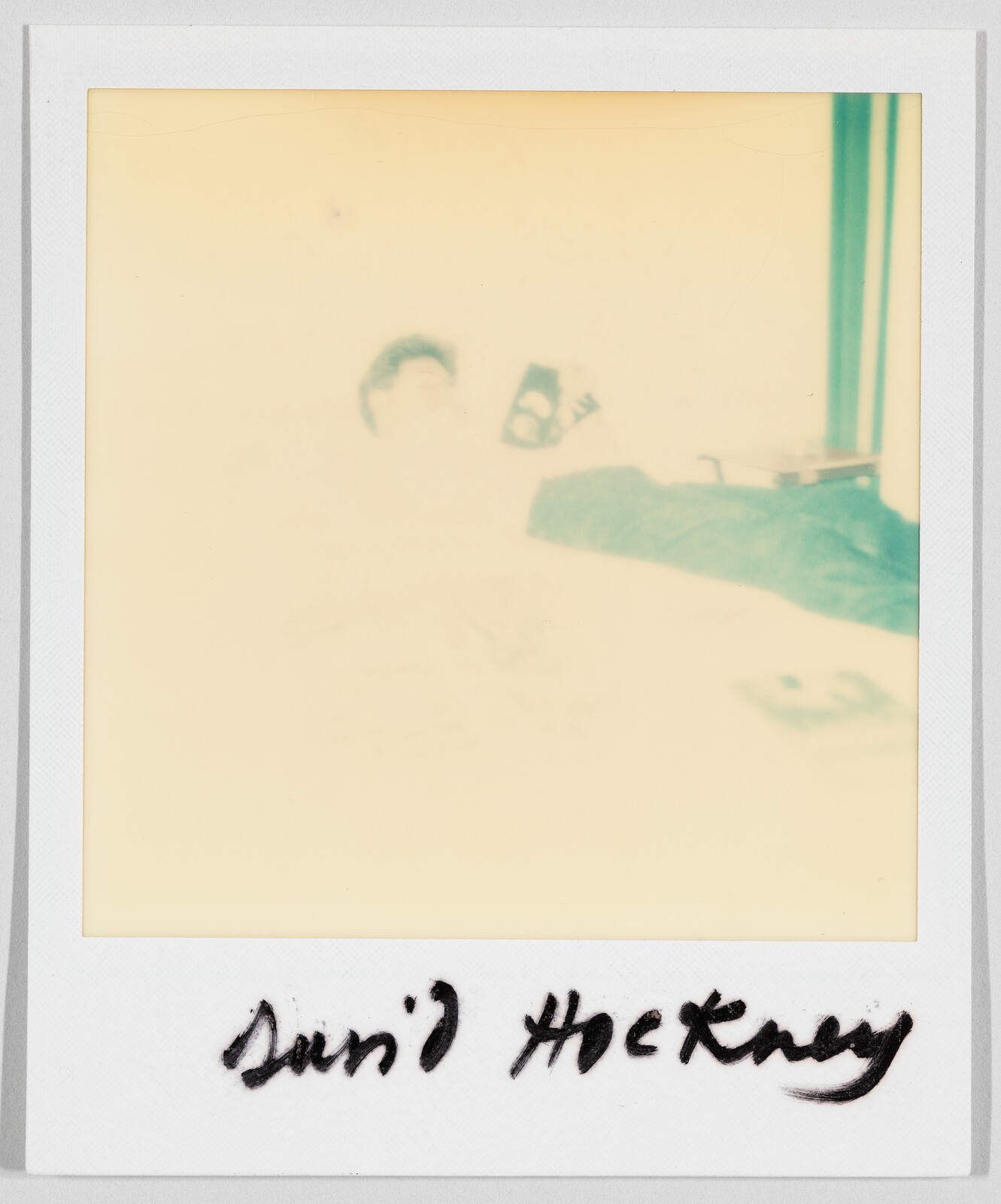 Faded Polaroid showing a person lying down, with handwritten text below reading “David Hockney.”