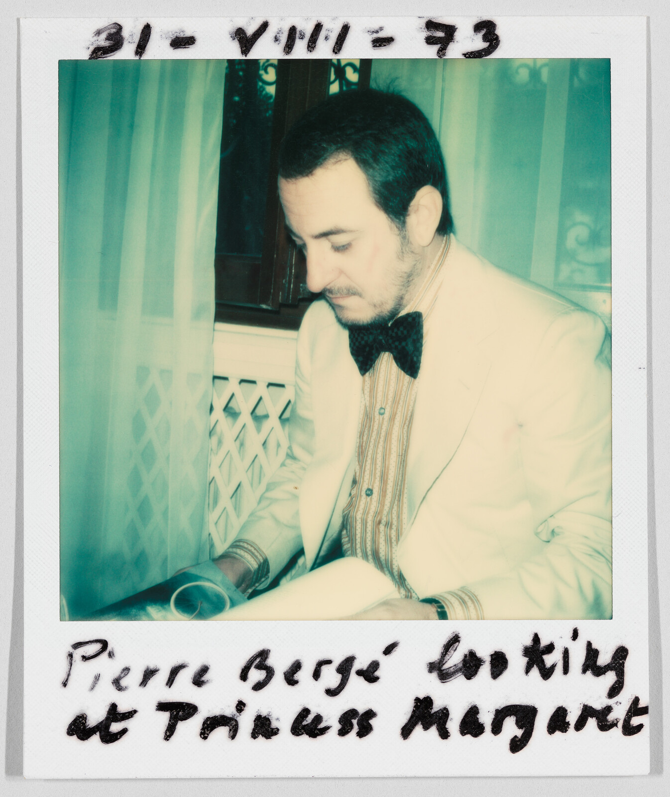 A man in a suit and bow tie sits and looks down at a magazine. Handwritten text reads: "Pierre Bergé looking at Princess Margaret."