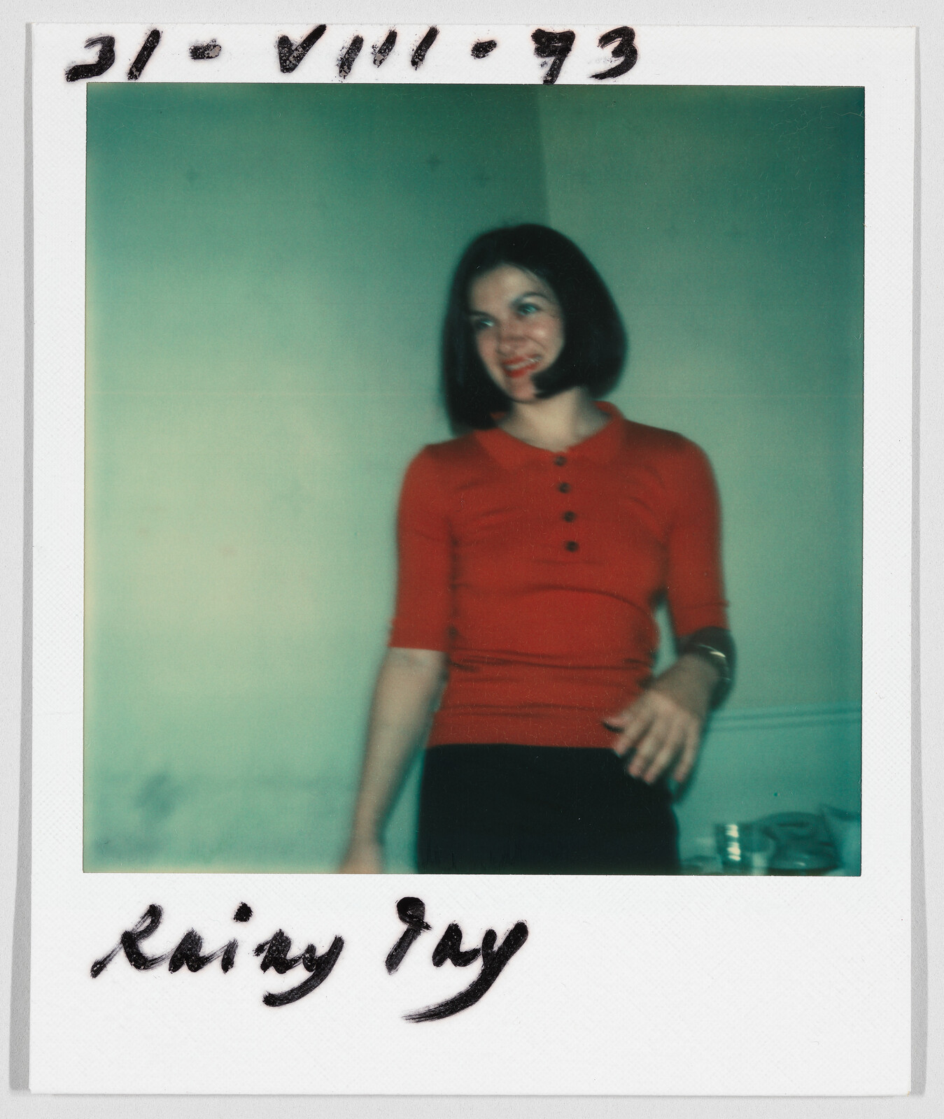 Woman with short dark hair wearing a red shirt, smiling and standing indoors. Handwritten date and name visible.