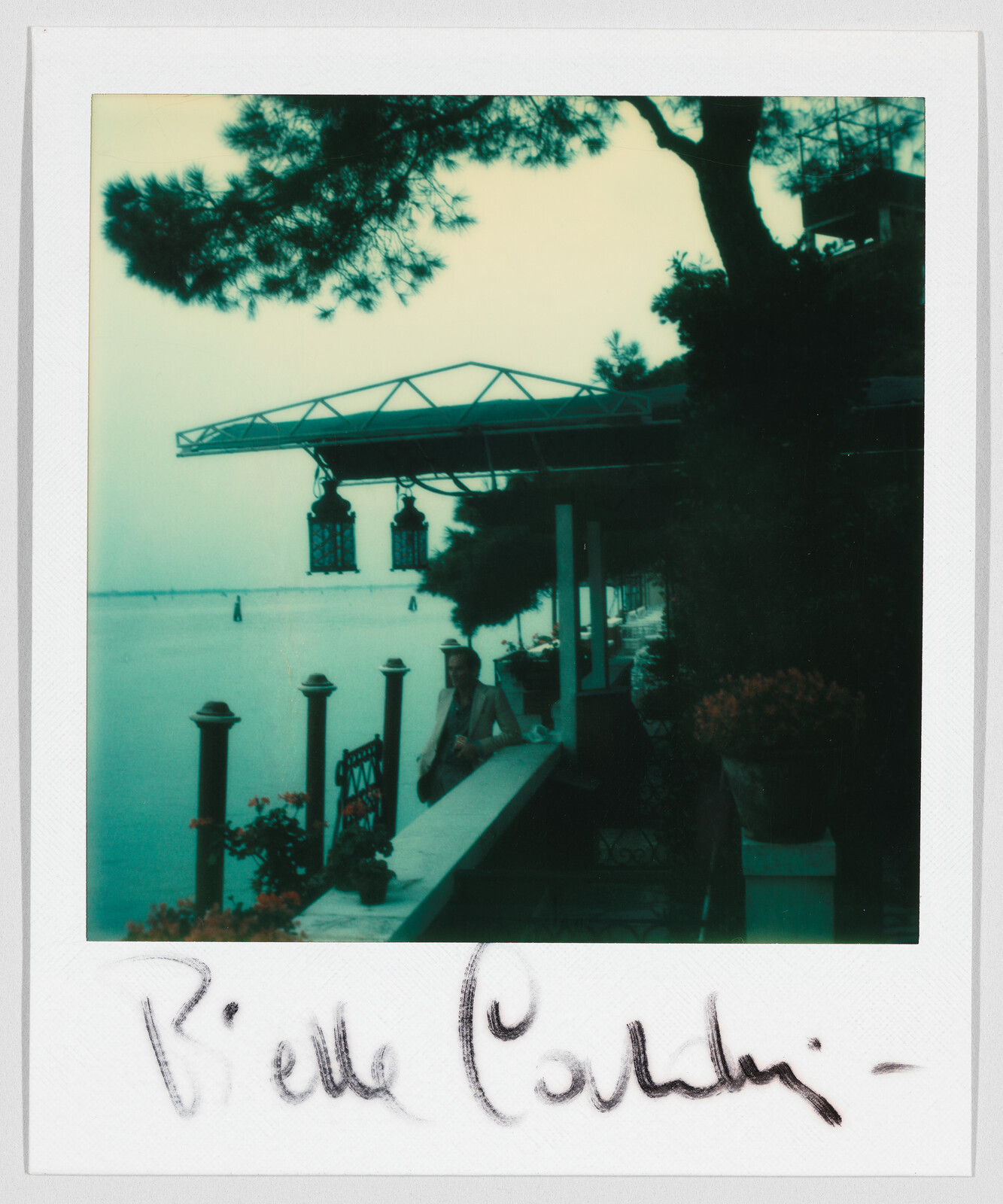 Person sitting by a waterfront patio with trees and hanging lanterns, handwritten text “Biella Cardin” at the bottom.