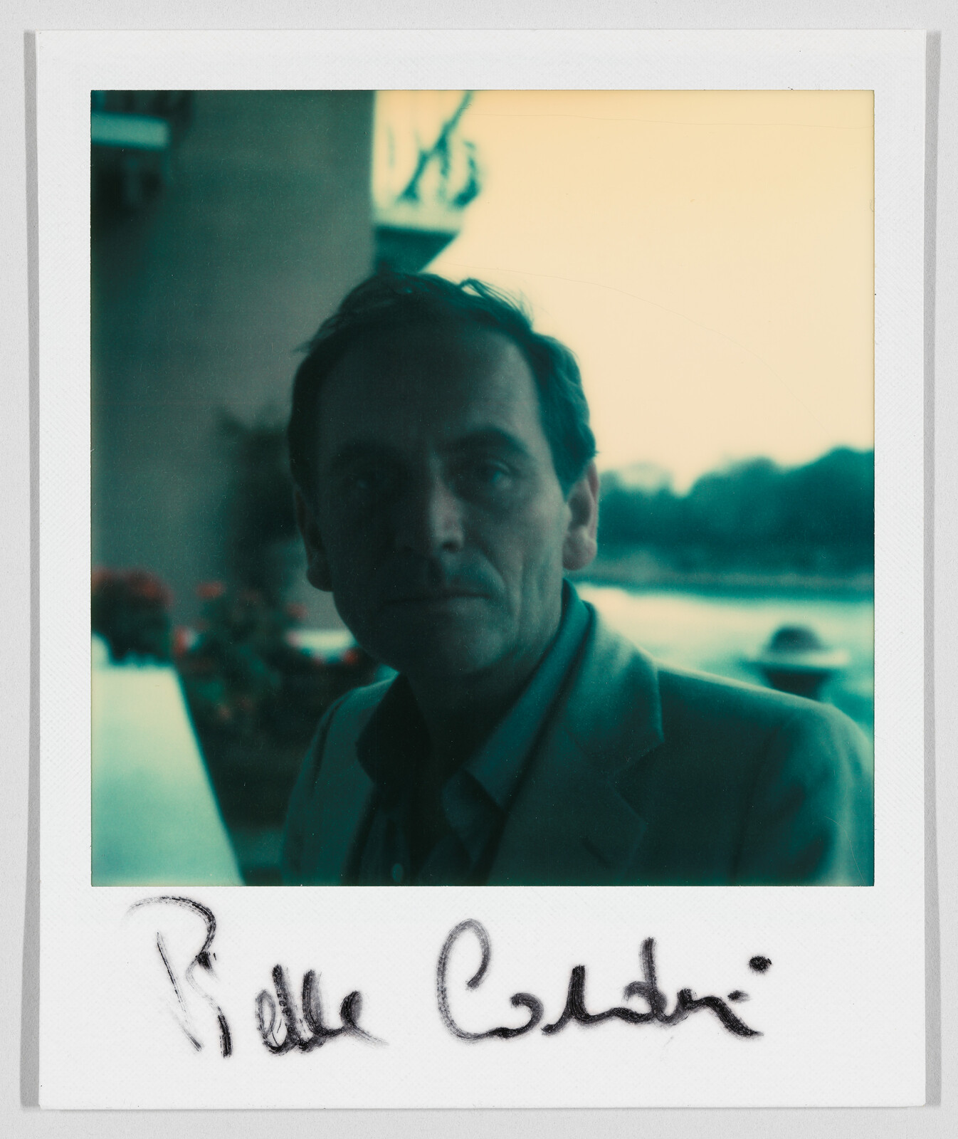 A man in a suit stands outdoors near water, with the handwritten name "Pierre Cardin" below.