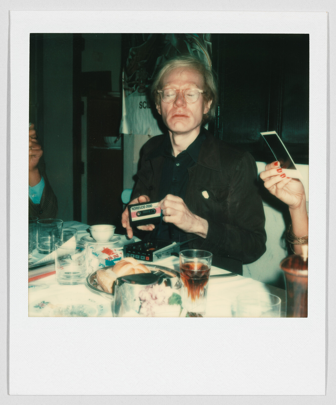 A man with glasses sits at a table holding a box labeled "NORELCO 200" among food and drinks.