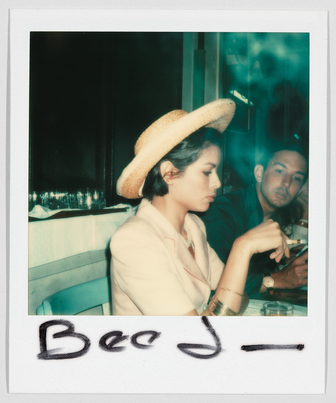 Two people sit at a table; the woman in front wears a straw hat and holds a cigarette. Handwritten text below reads "Bee J_".