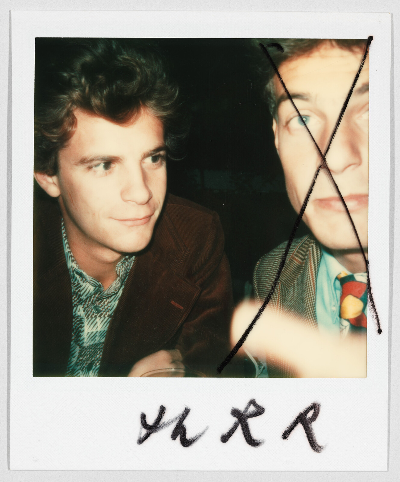 Two men sitting together; the man on the right has a large black X drawn over his face.