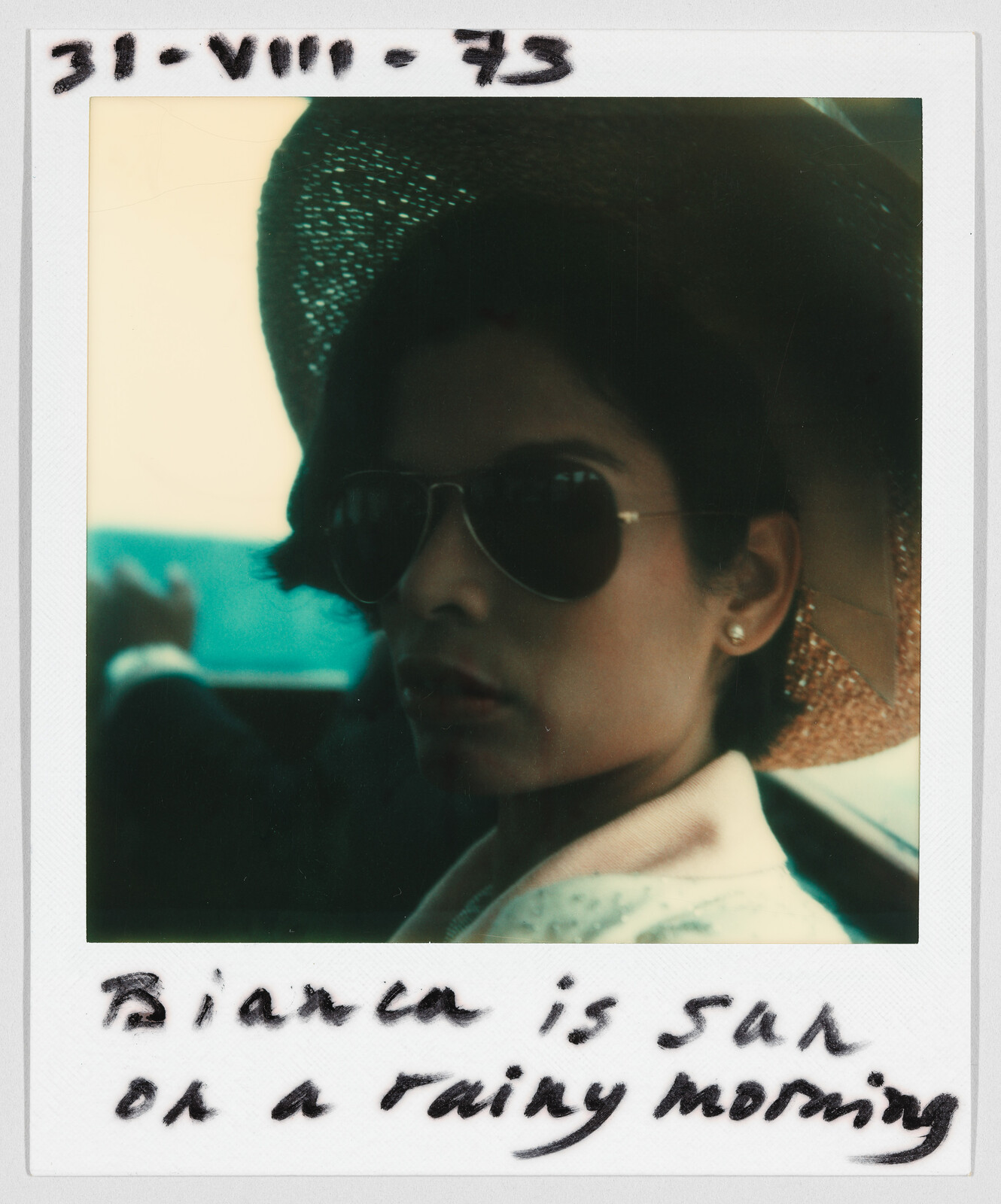 Woman wearing sunglasses and a hat sits in a car. Handwritten text says, "Bianca is sad on a rainy morning."