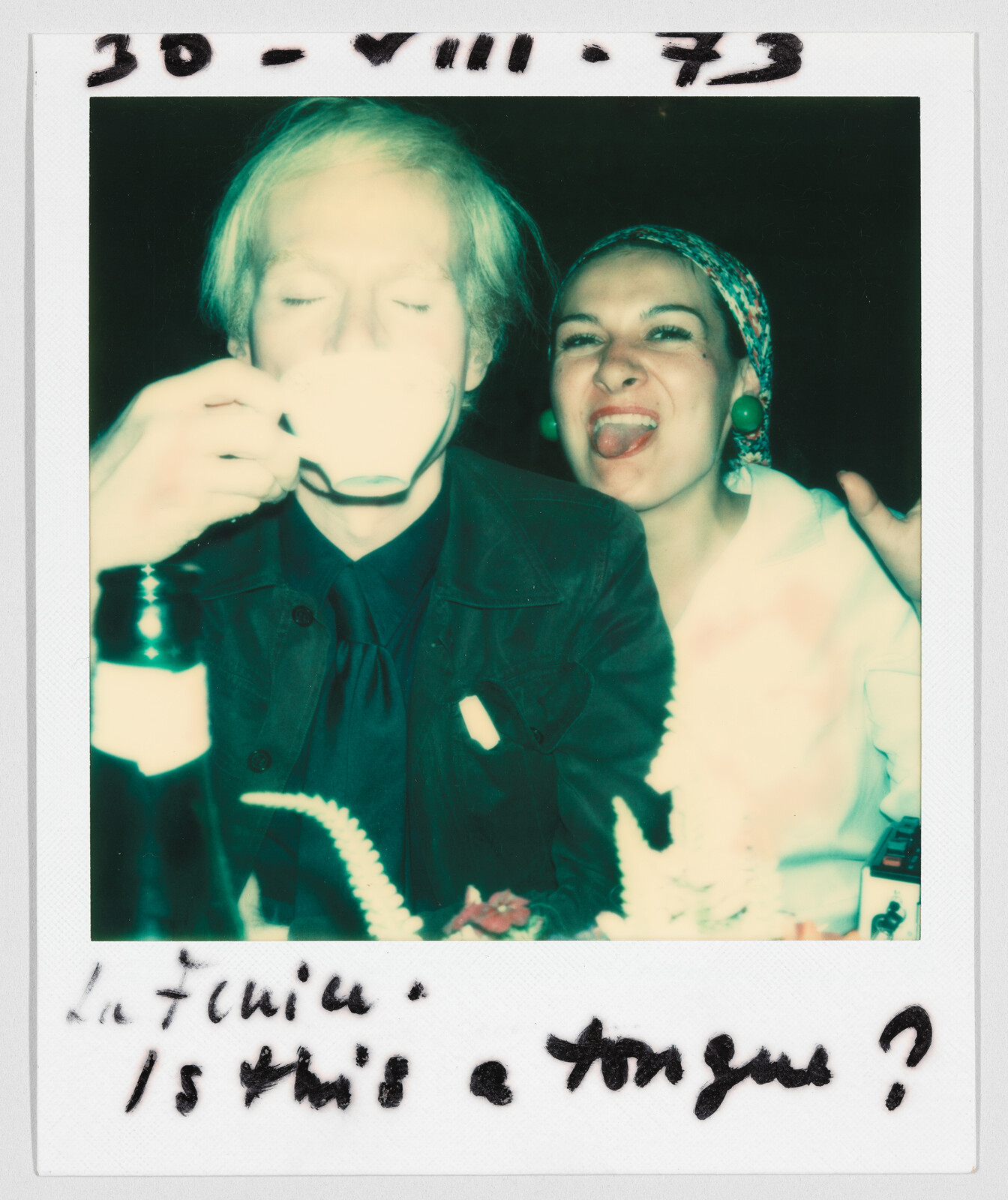 A man drinks from a teacup while a woman next to him sticks out her tongue and smiles. Handwritten text reads, “Is this a tongue?”
