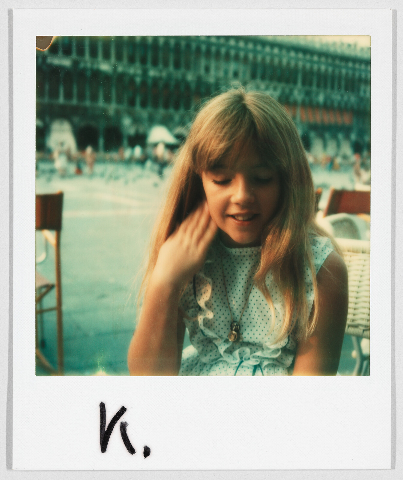 Young girl with long blonde hair sits outdoors, touching her hair. The letter "K." is written below.