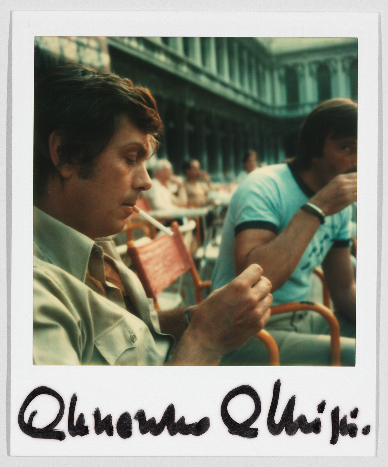 Two men sit at an outdoor café; one is lighting a cigarette, the other is drinking from a cup.