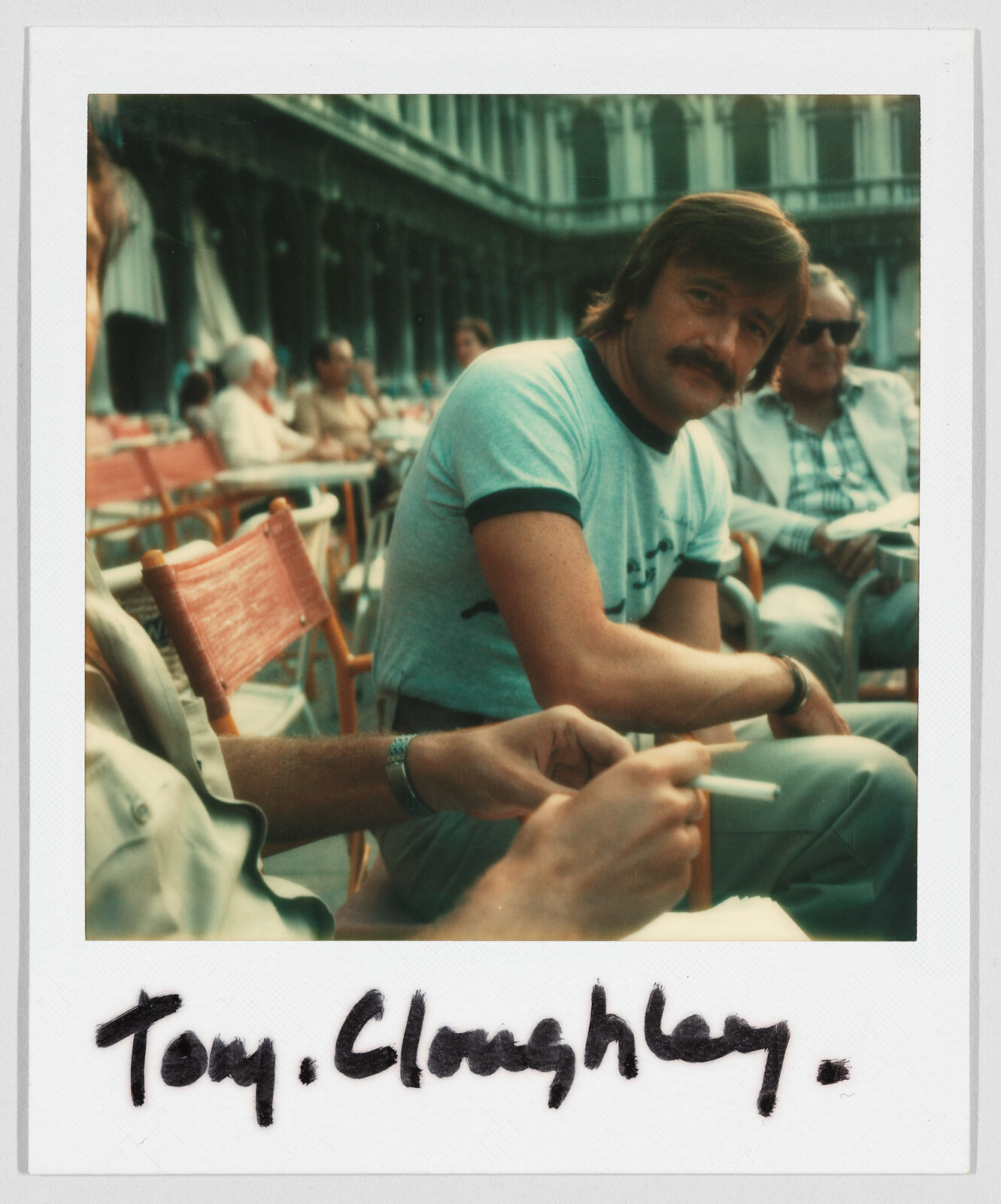 Man with a mustache sits at an outdoor café table; handwritten text below reads “Tony Cloughley.”