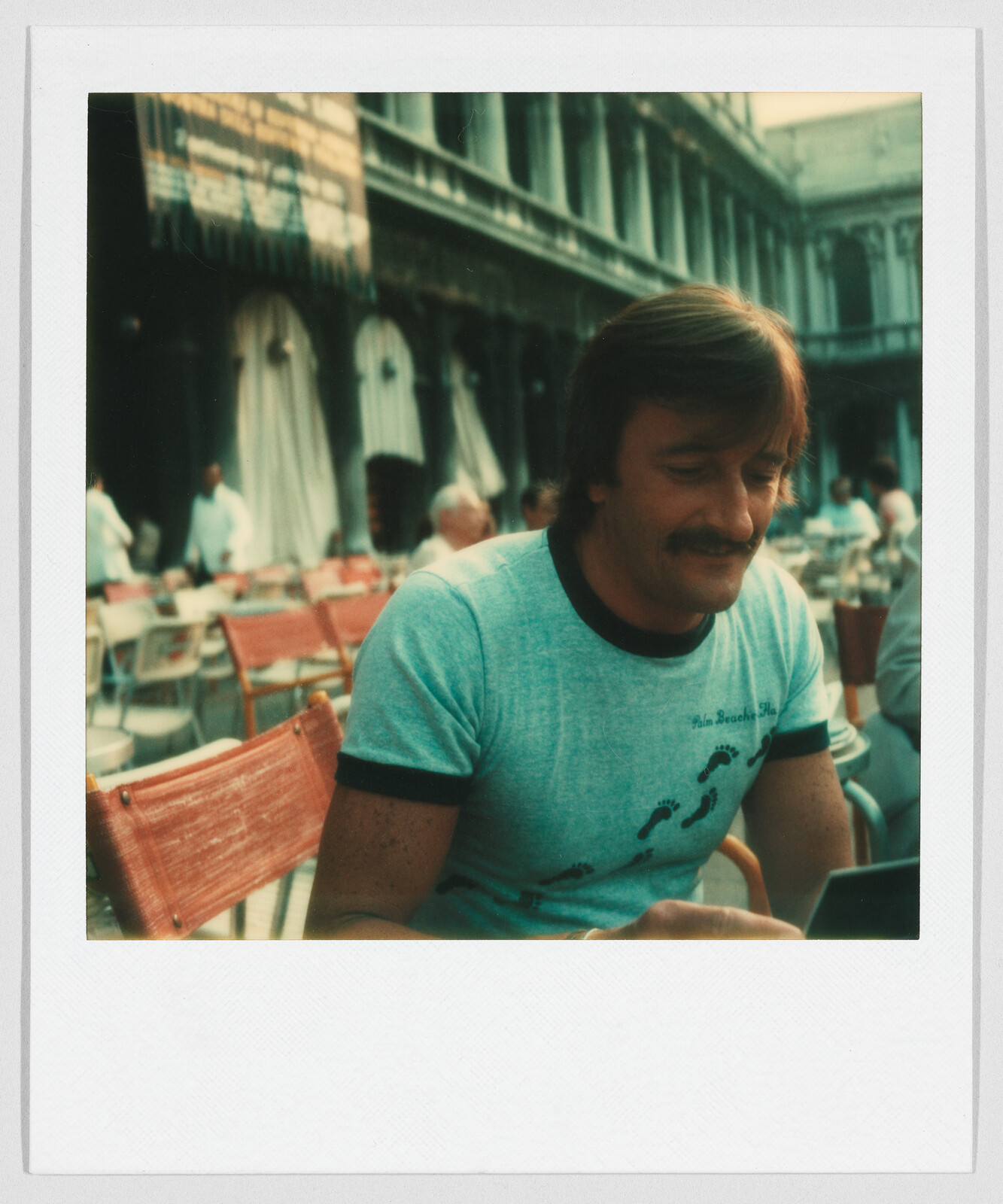 Man with a mustache sits at an outdoor café, wearing a light blue t-shirt with black footprints.