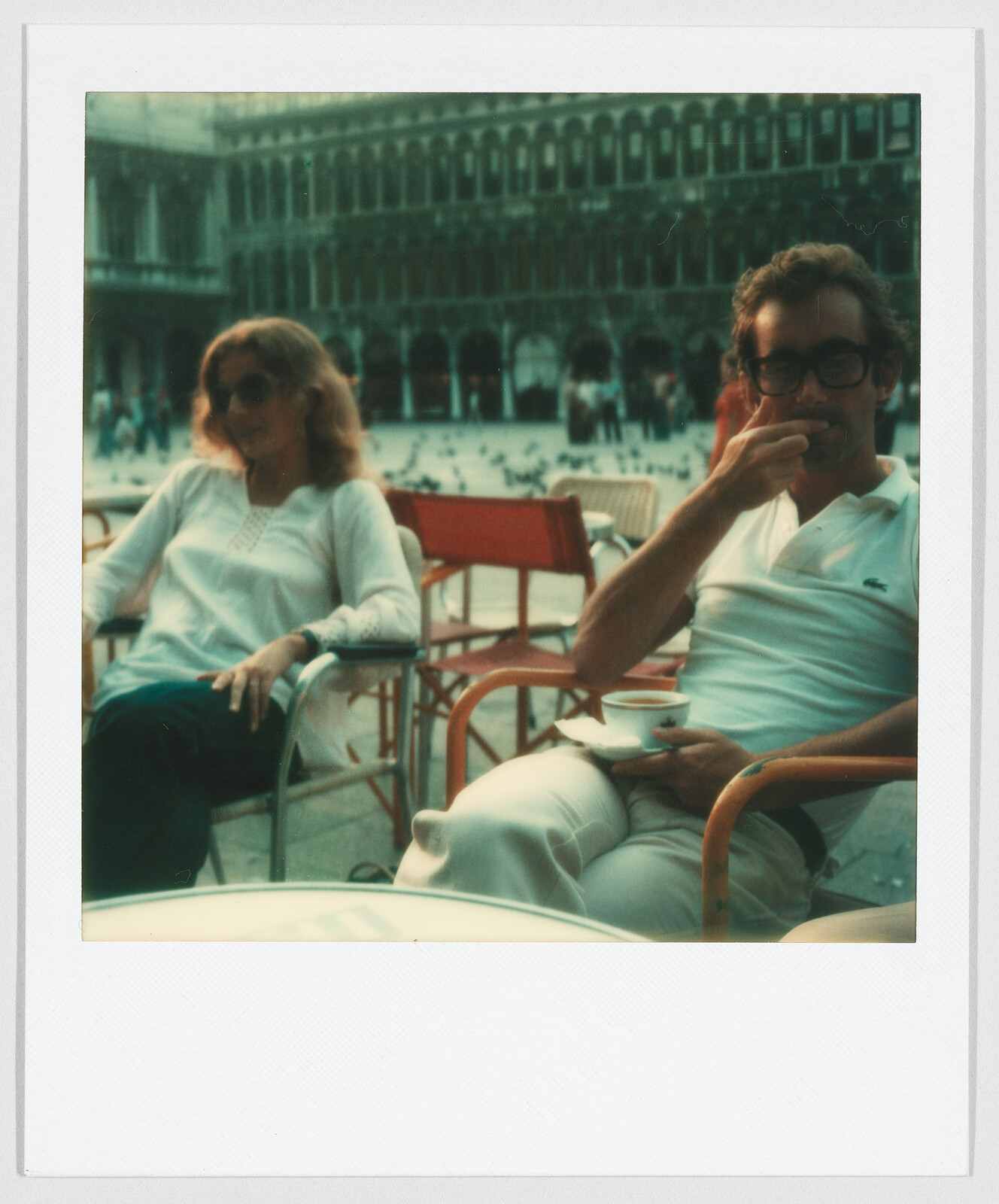 Two people sit at an outdoor café, one drinking from a cup, with a large building and pigeons in the background.