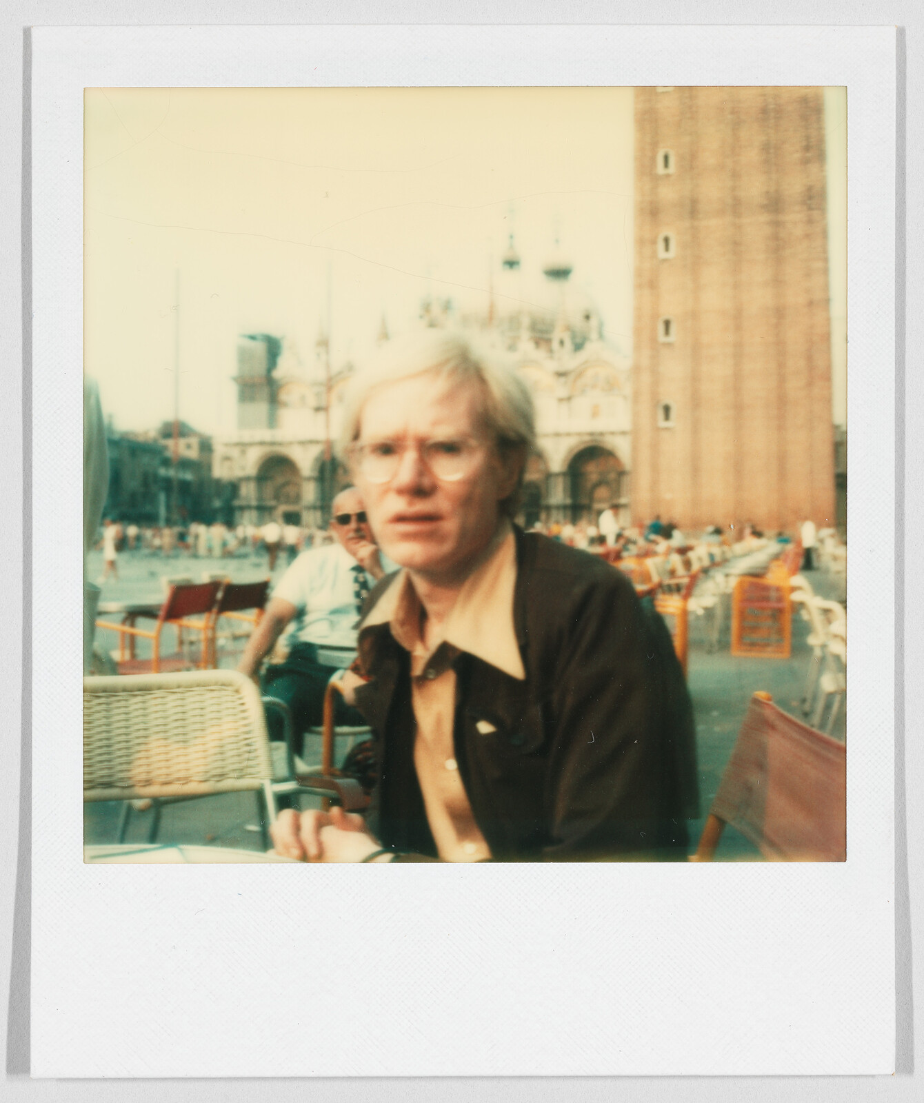 A man with light hair and glasses sits at an outdoor café with historic buildings in the background.