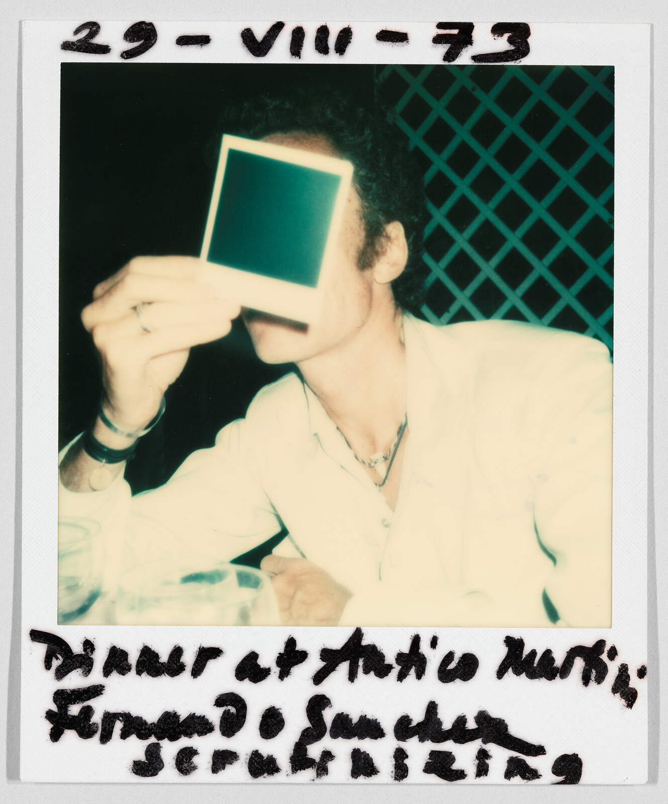 Person in a white jacket holds a blank photo in front of their face; handwritten date and notes visible.