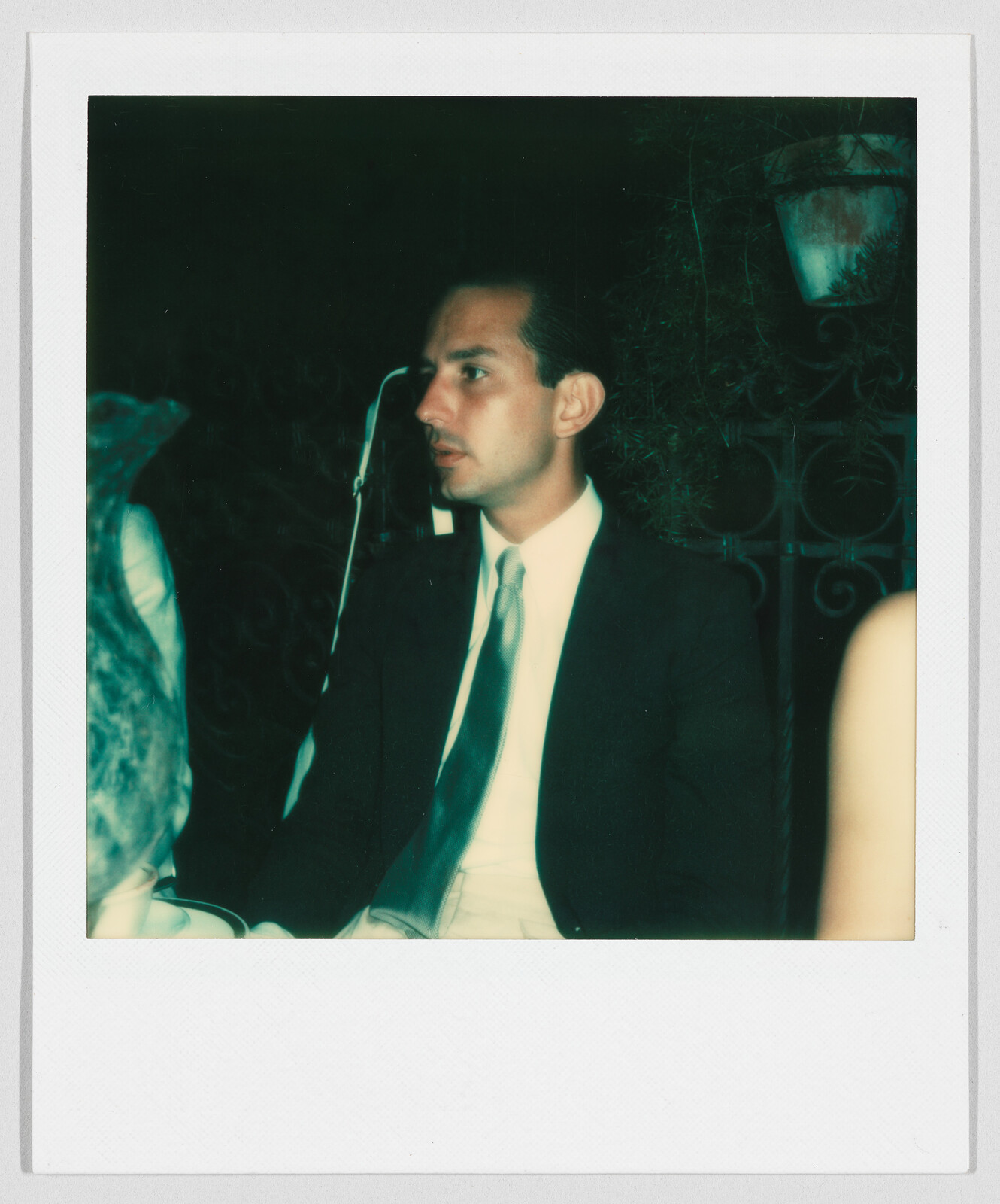 A man in a suit and tie sits at a table, looking to the side in a dimly lit setting.