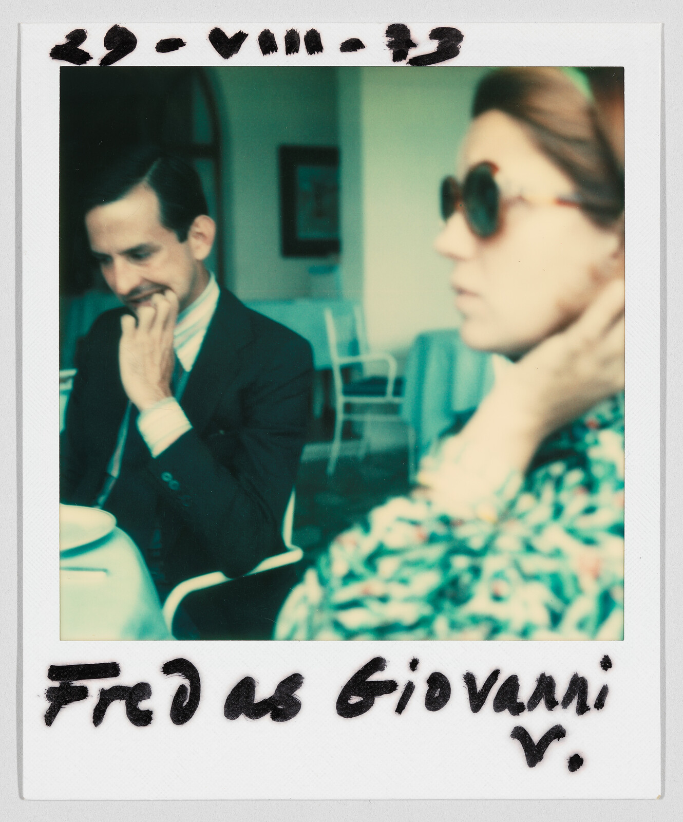 A man in a suit sits at a table, while a woman in sunglasses touches her neck. Text reads: "Fred as Giovanni."