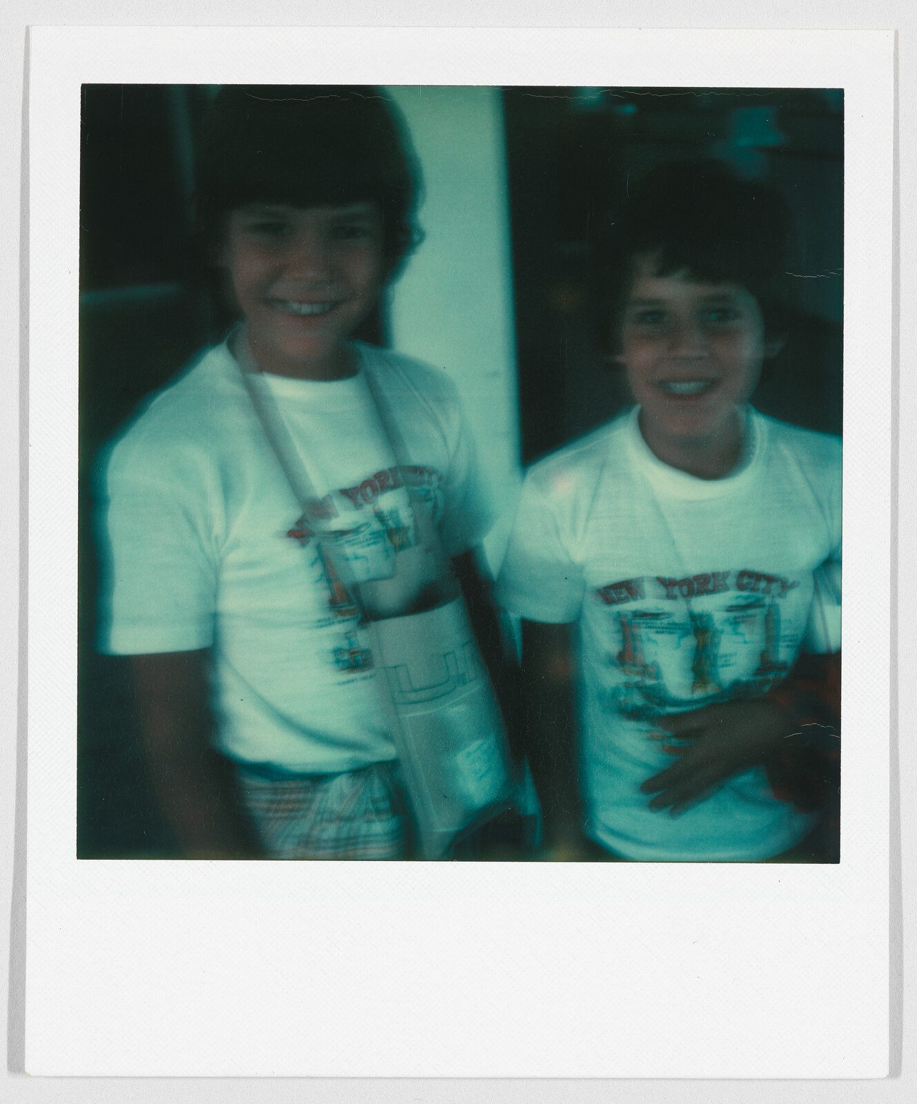 Two children smiling, both wearing white “New York City” t-shirts, standing close together indoors.