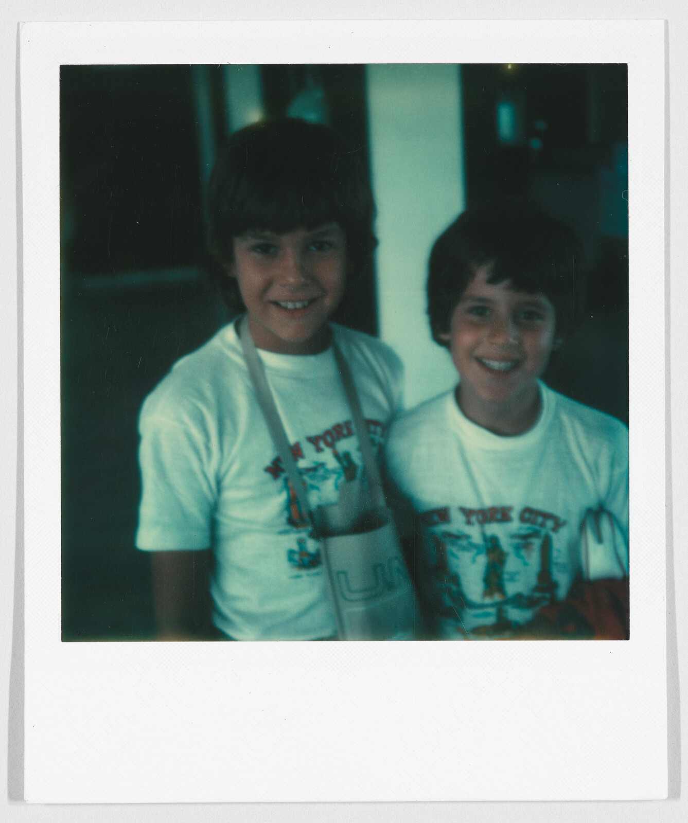 Two smiling children wearing matching “New York City” t-shirts stand close together, one holding a bag.