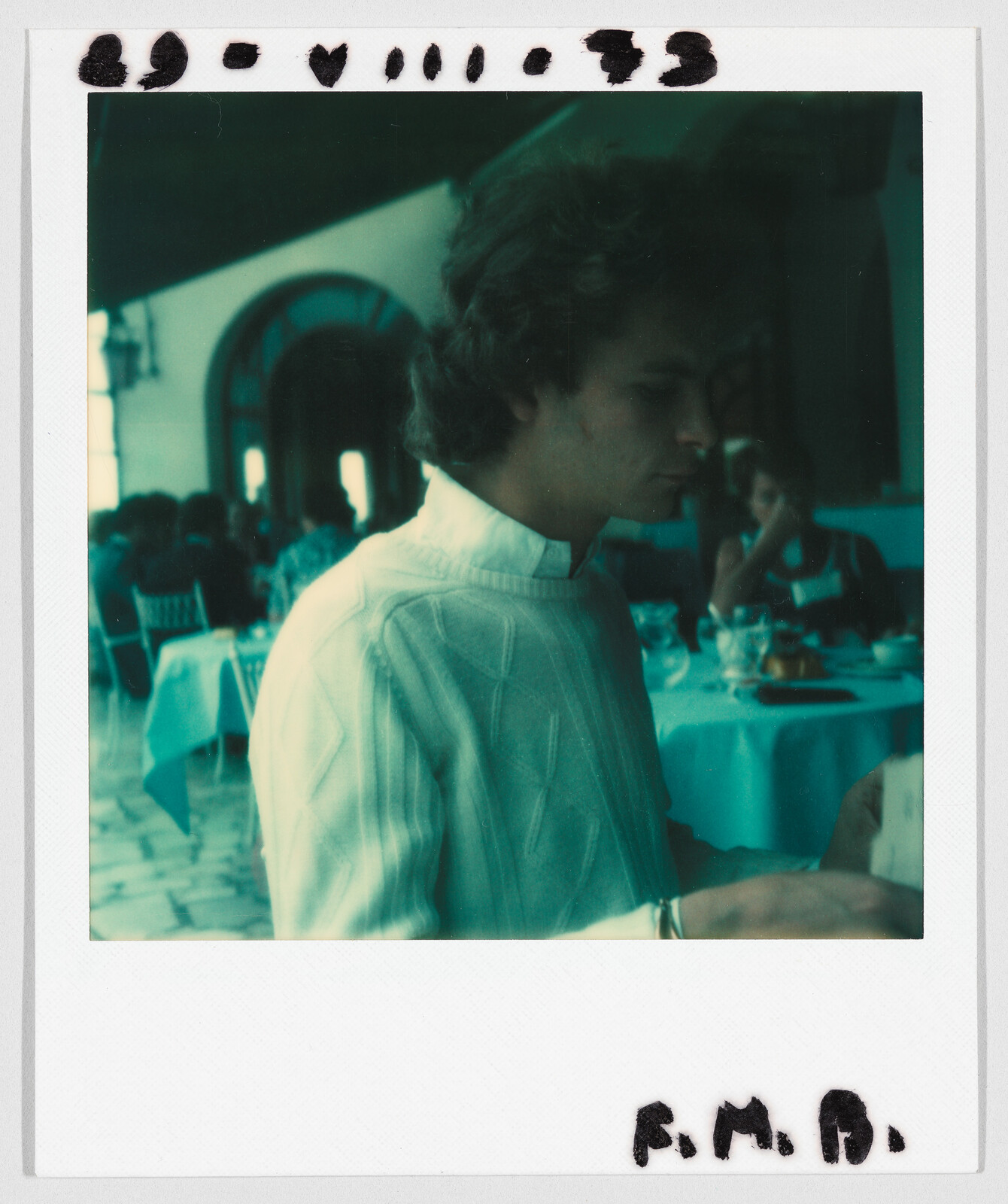 A young person in a white shirt sits at a table in a restaurant, looking down. Handwritten text: "F.M.B."